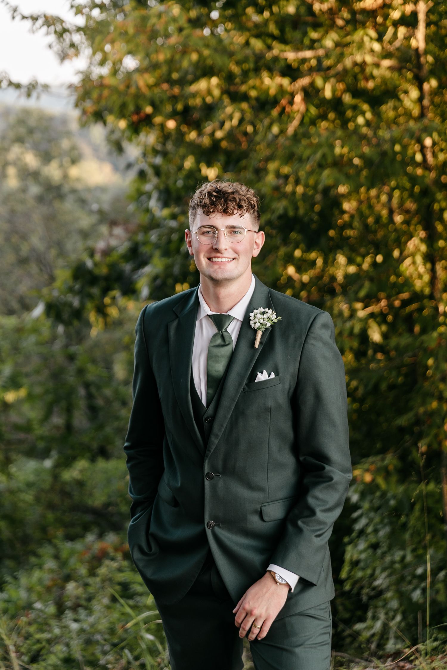 A groom in a black suit poses outdoors against autumn foliage in a series of wedding portraits.