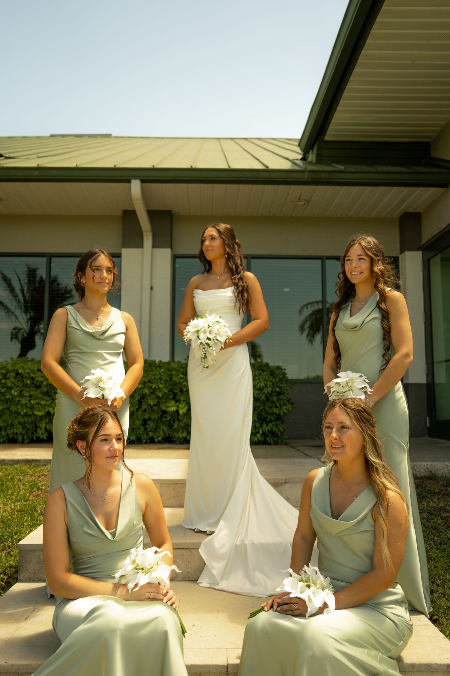 Bride in white gown holds bouquet flanked by four bridesmaids in sage green dresses outside a building.