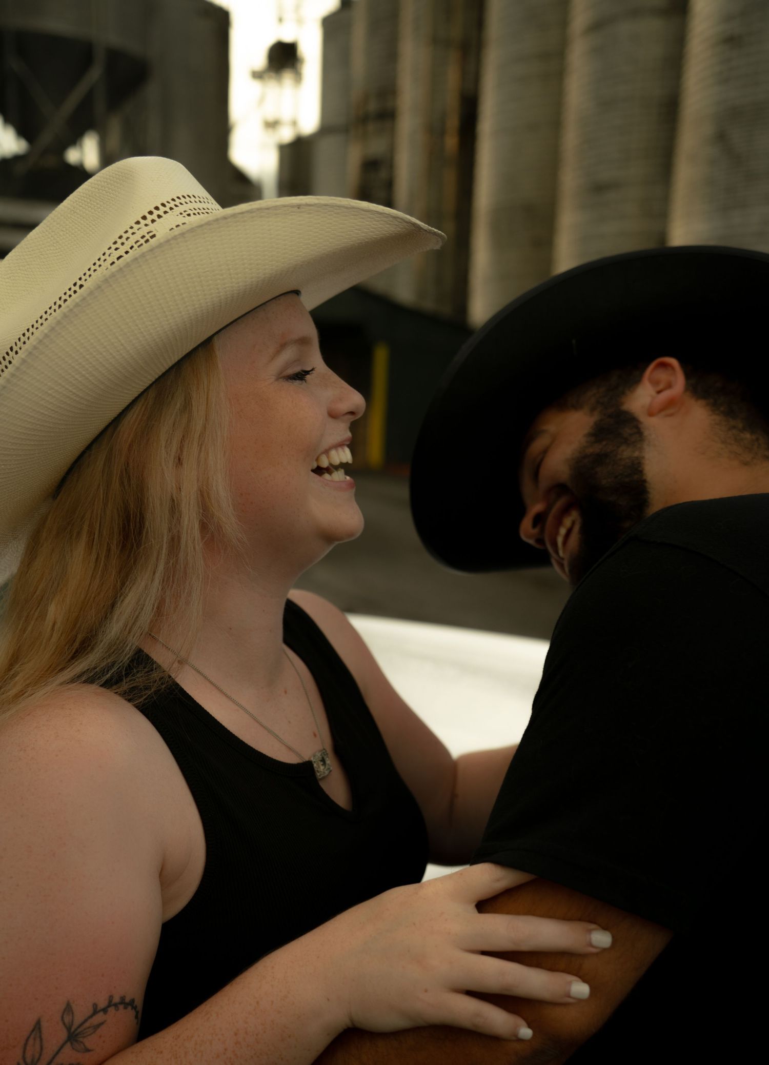 A smiling woman in a white cowboy hat embraces a man in a black cowboy hat outdoors near industrial silos.