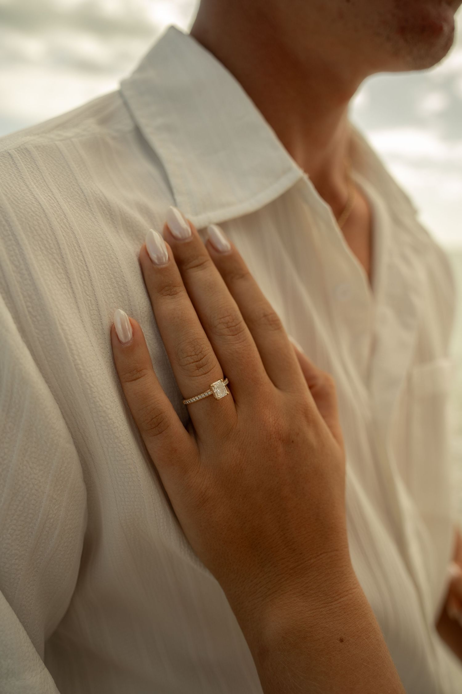 Close-up of a womans hand displaying a gold solitaire engagement ring resting on a mans white linen shirt.