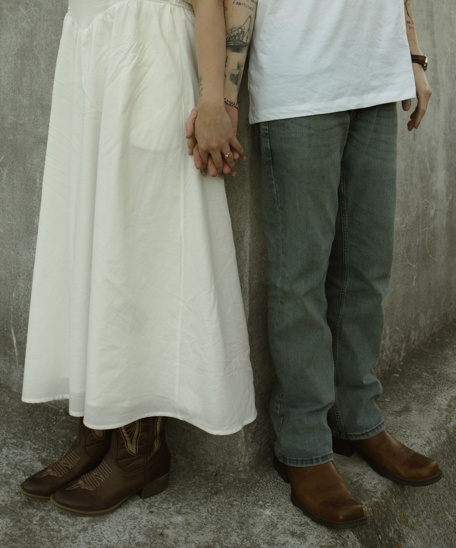 Couple holding hands wearing matching brown cowboy boots, white maxi skirt and gray jeans, standing against a concrete wall.