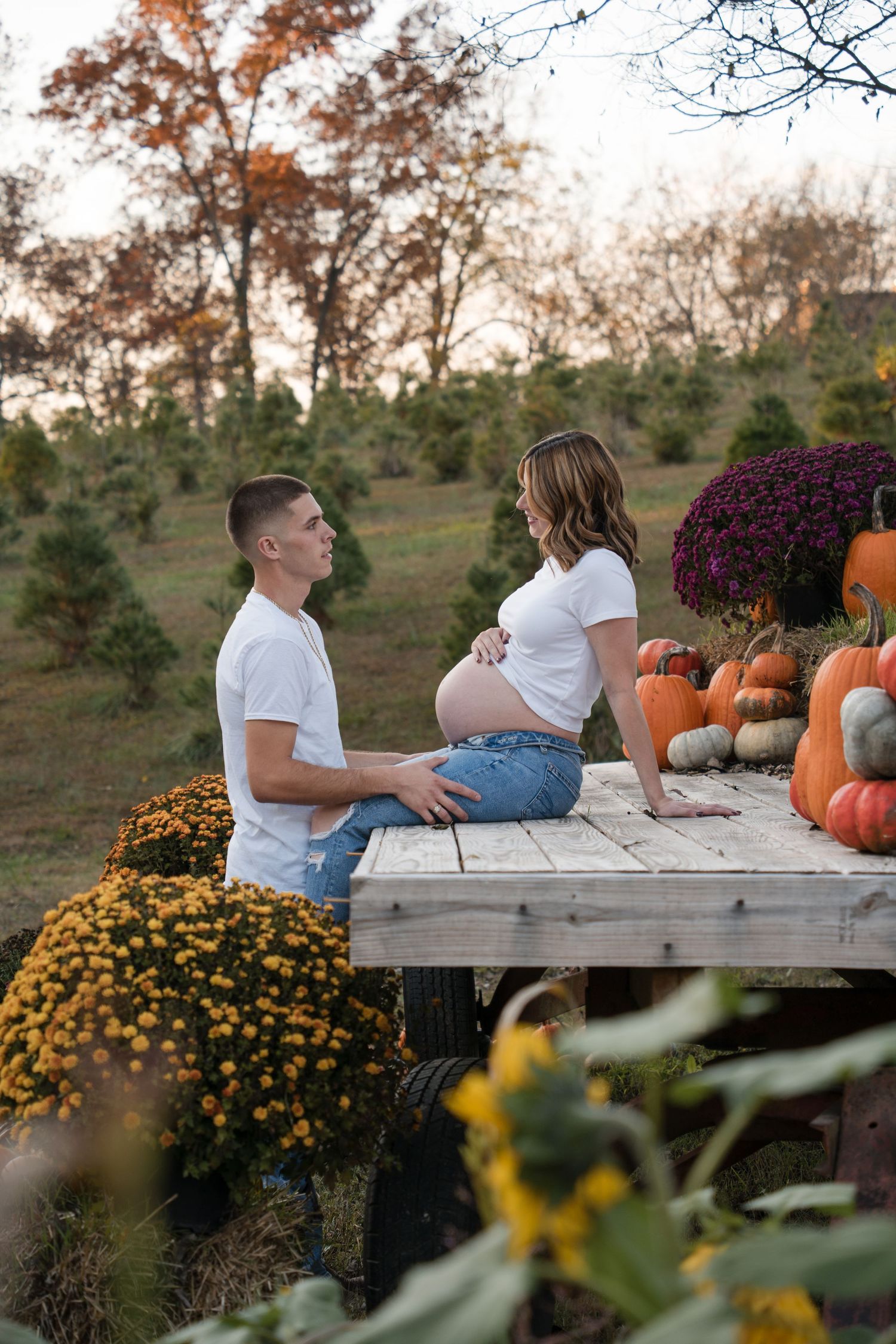 A couple lounges on a wooden bench surrounded by autumn pumpkins and yellow mums in a garden setting.