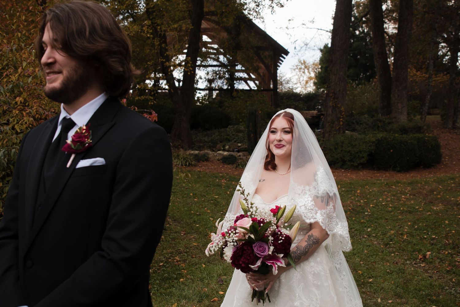 A couple in wedding attire poses playfully in an autumn park setting with fallen leaves.
