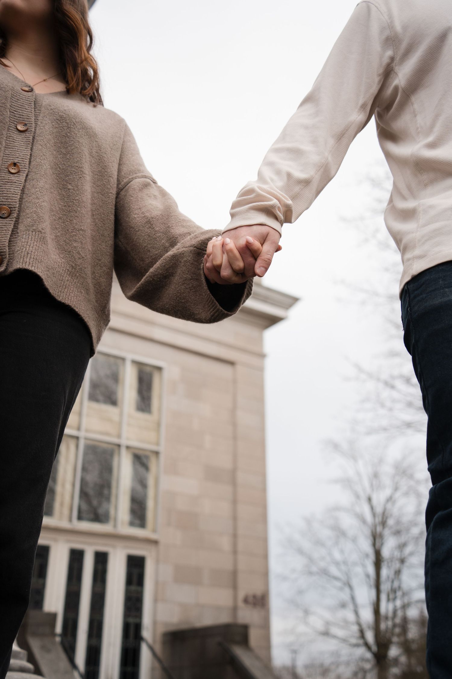 A couple in casual attire holding hands while walking outside a building on an overcast day.