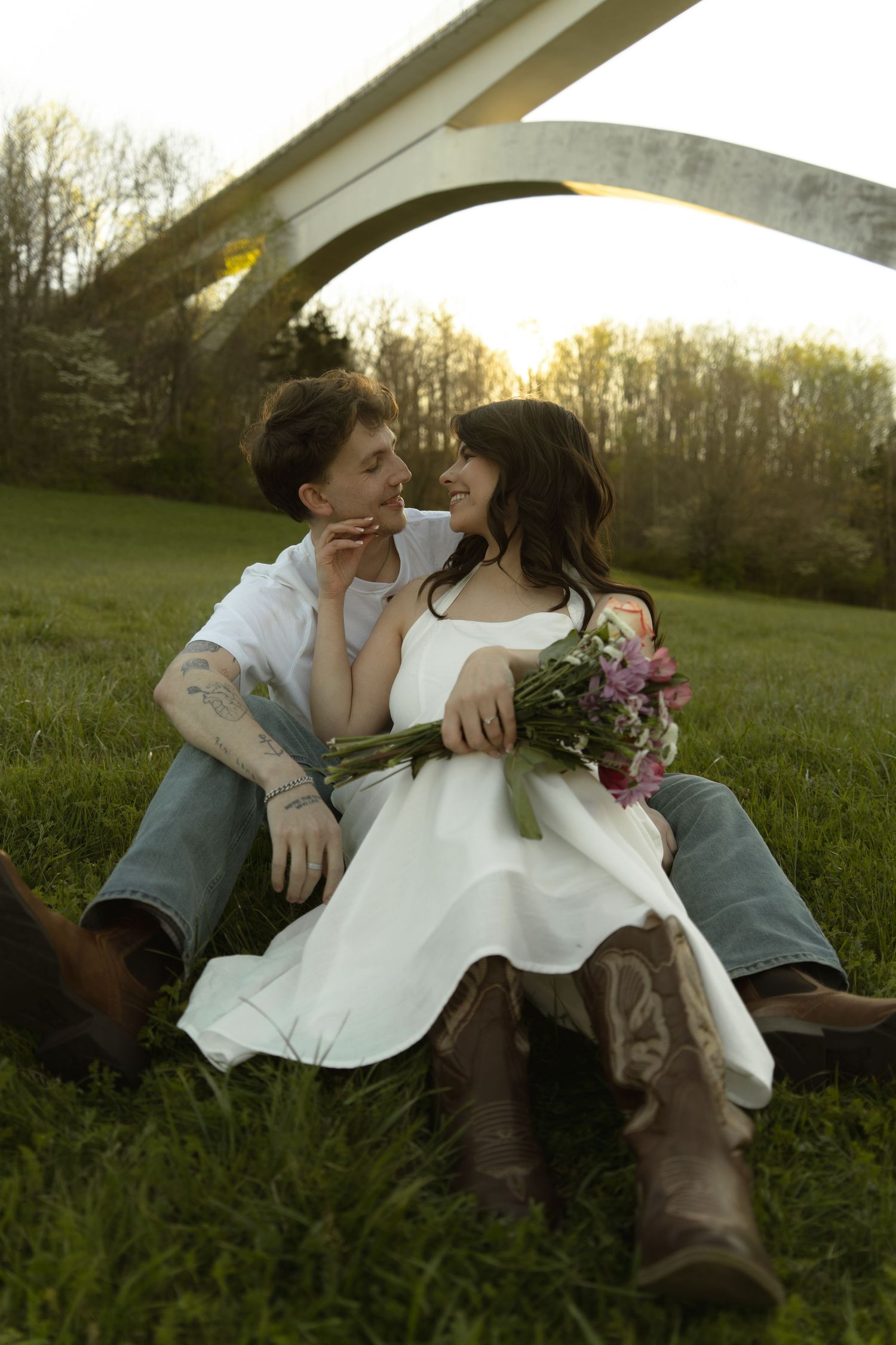A couple in wedding attire sits on green grass, gazing at each other, bride holding a pink bouquet near an arch.
