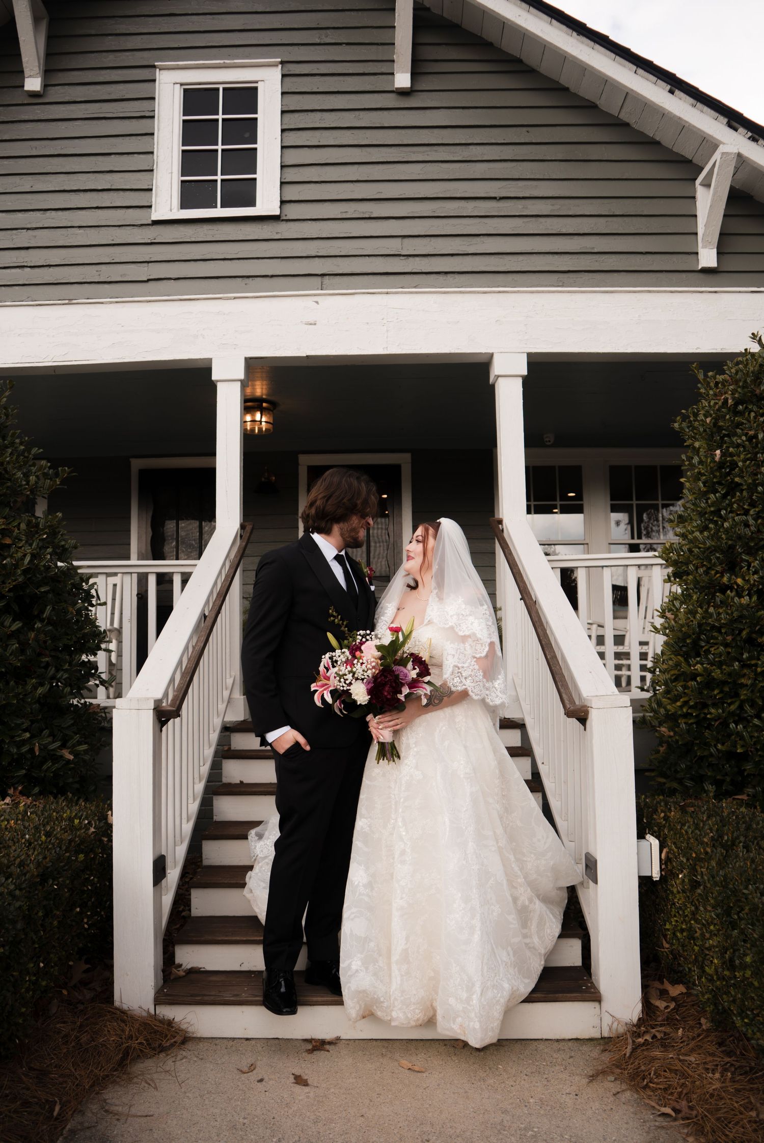 A couple shares a romantic moment on a white porch of a gray house during a wedding photoshoot.