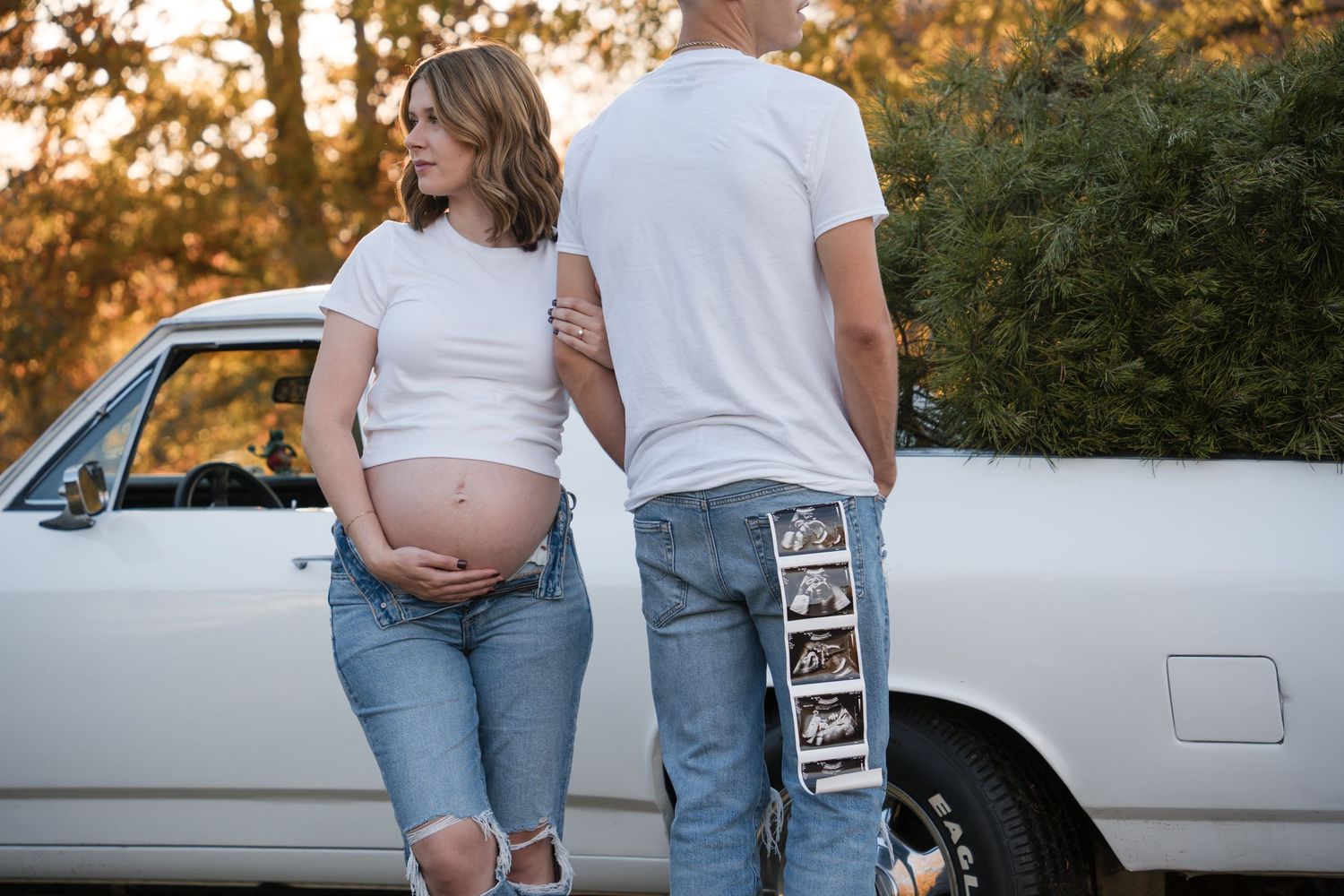 Expecting couple stands beside vintage car at sunset holding ultrasound photos in rustic outdoor setting.