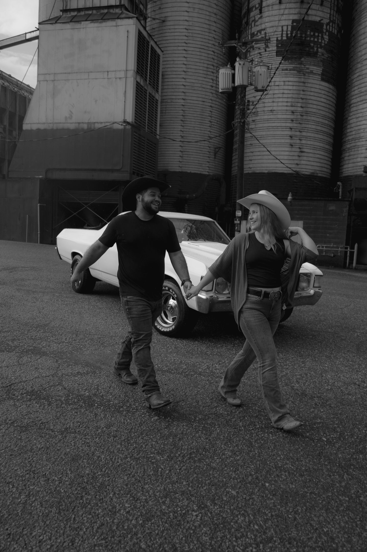 Black and white photo of a couple in cowboy hats holding hands near a classic white car and grain silos.