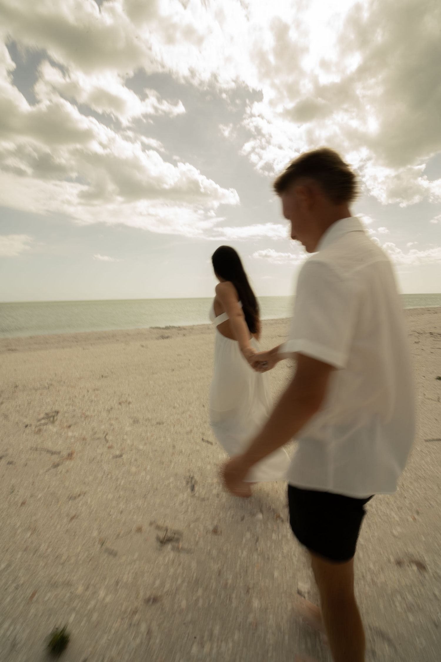Couple in white walking on a sandy beach with dramatic cloudy sky and ocean in the background.