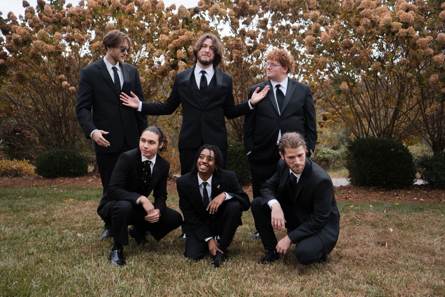 A wedding party poses playfully in formal attire among autumn trees and fallen leaves.