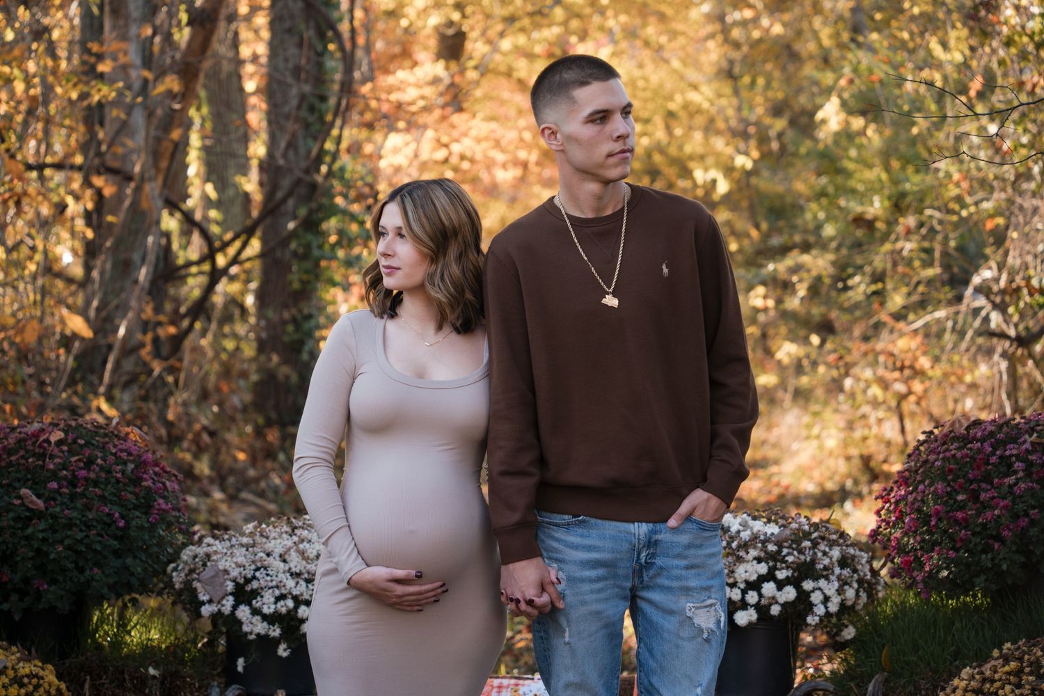 A couple stands together in an autumn garden setting with warm golden foliage and white flowers in the background.