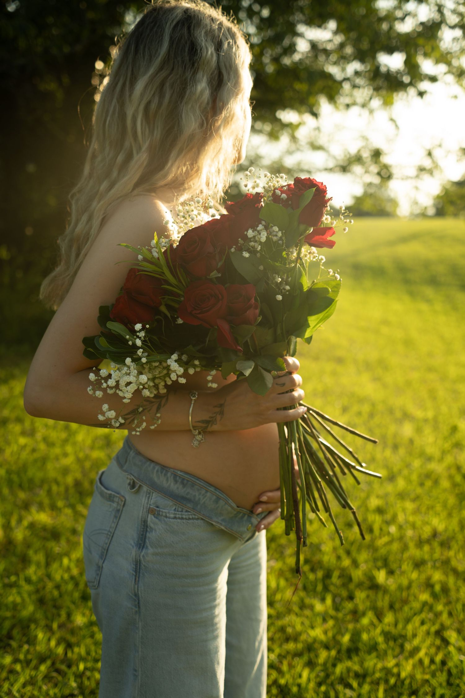 Blonde woman holding red roses bouquet in sunny green field, wearing low-rise jeans, golden hour light.