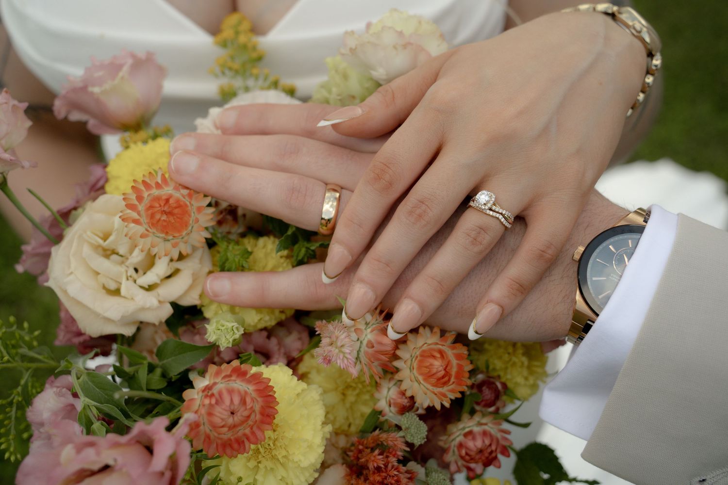 Newlywed couple displaying wedding rings on hands resting over a colorful bridal bouquet with pink and yellow flowers.