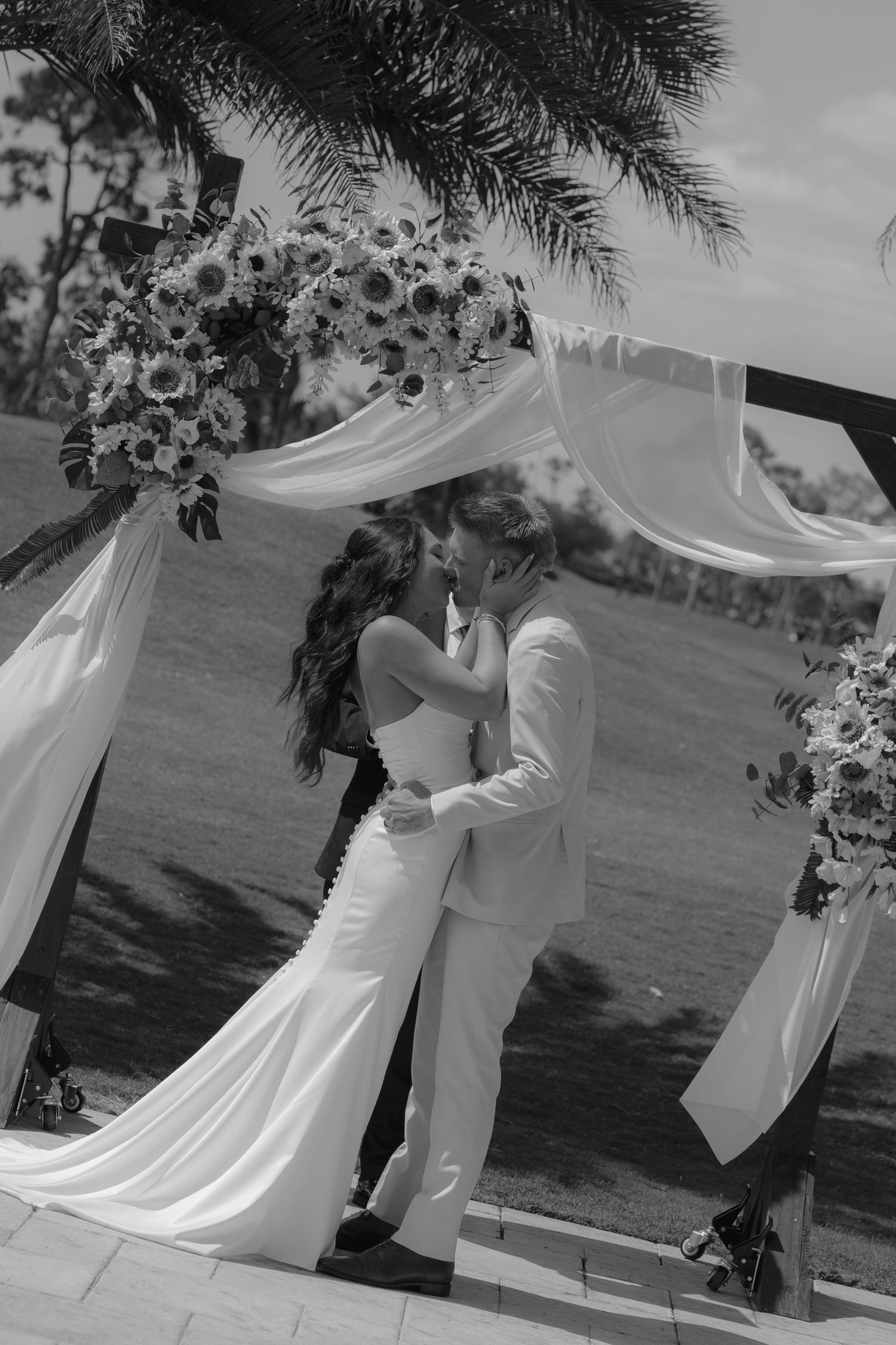 Black and white photo of a bride and groom sharing their first kiss under a floral arch draped with flowing fabric.