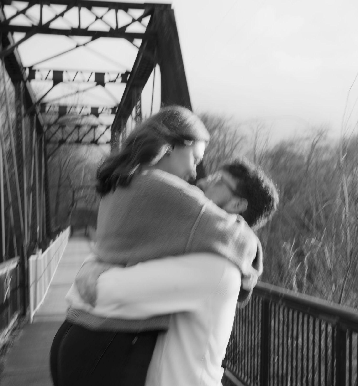 Black and white photo of a couple embracing on a bridge surrounded by trees.