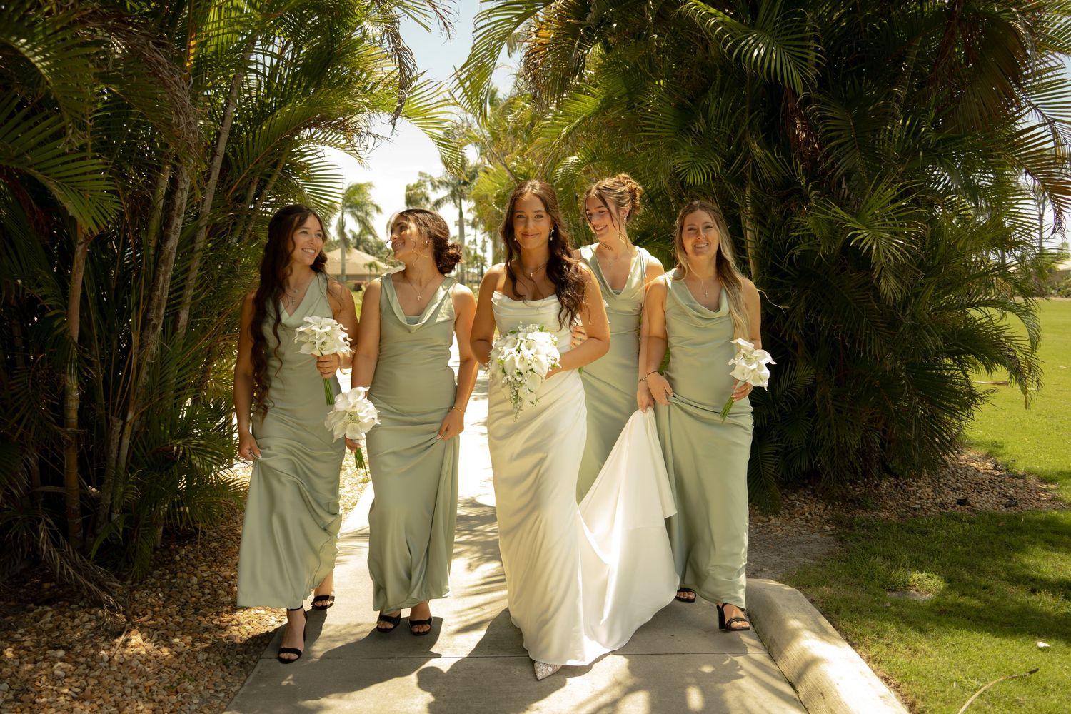 Bride in white gown walks with four bridesmaids in sage green dresses, holding white bouquets along a sunny tropical path.