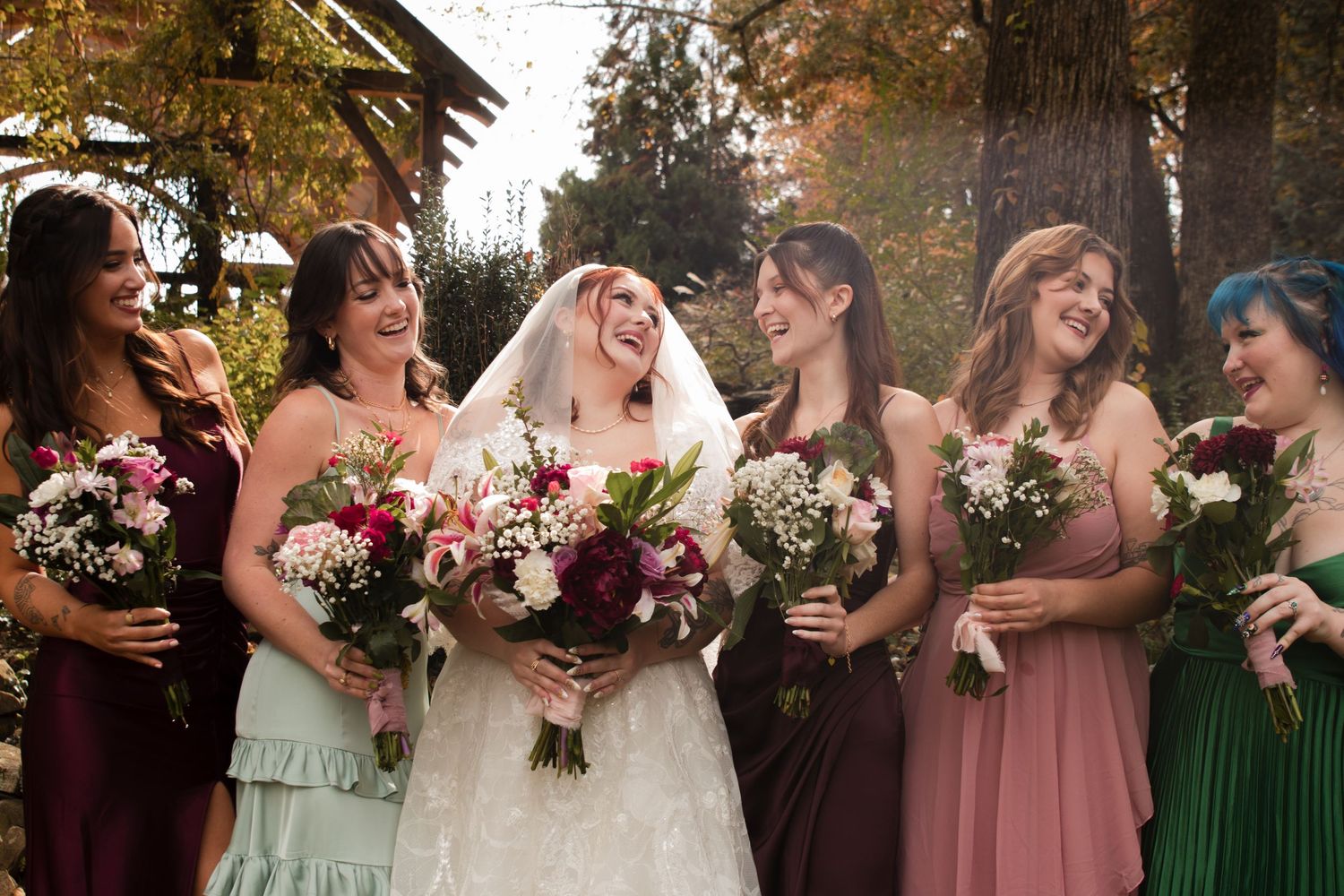 Wedding party members in burgundy, pink and green dresses pose together with bouquets during an autumn celebration.