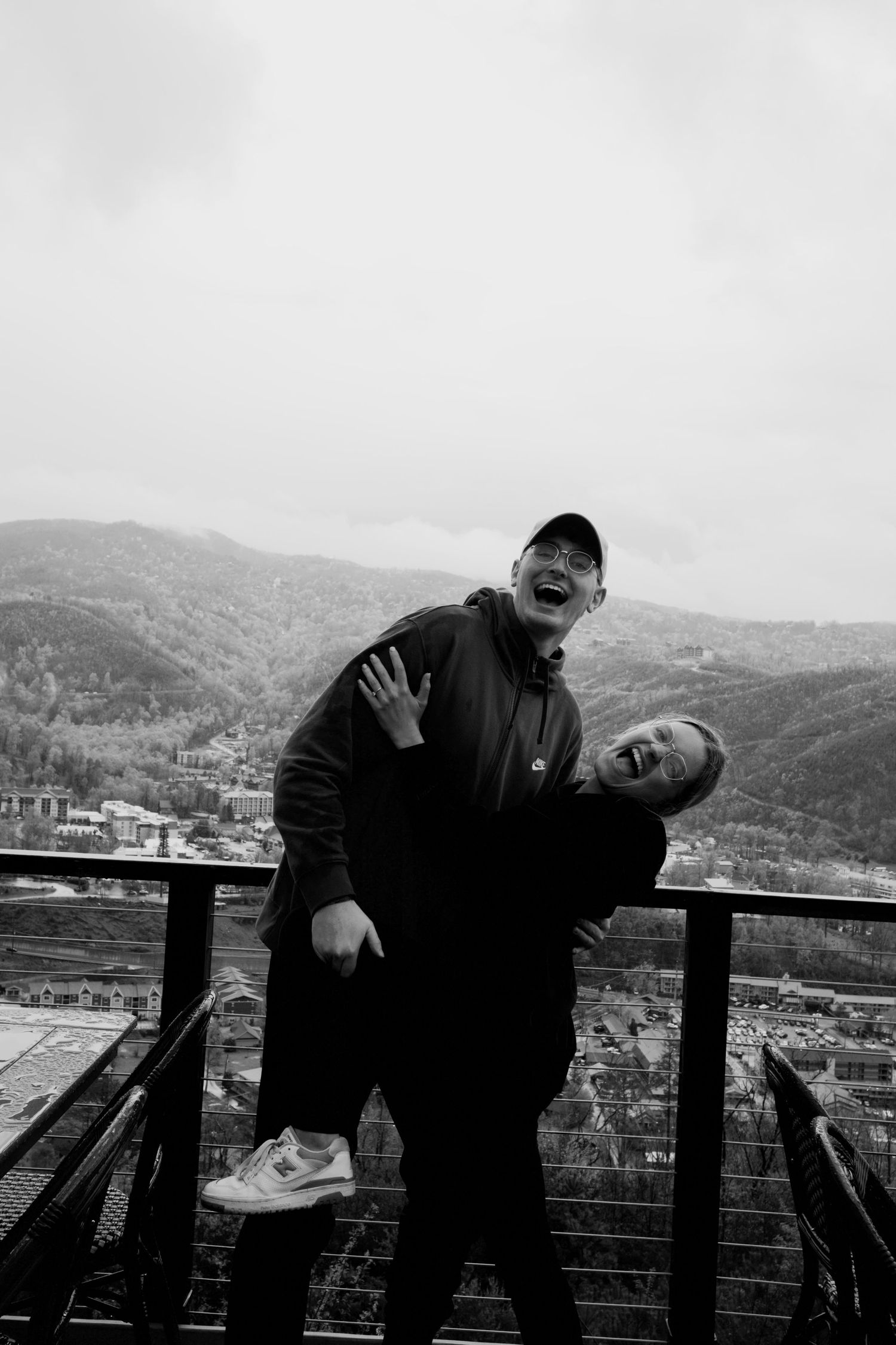Two people playfully posing and leaning over a railing with mountains in the background, captured in black and white.