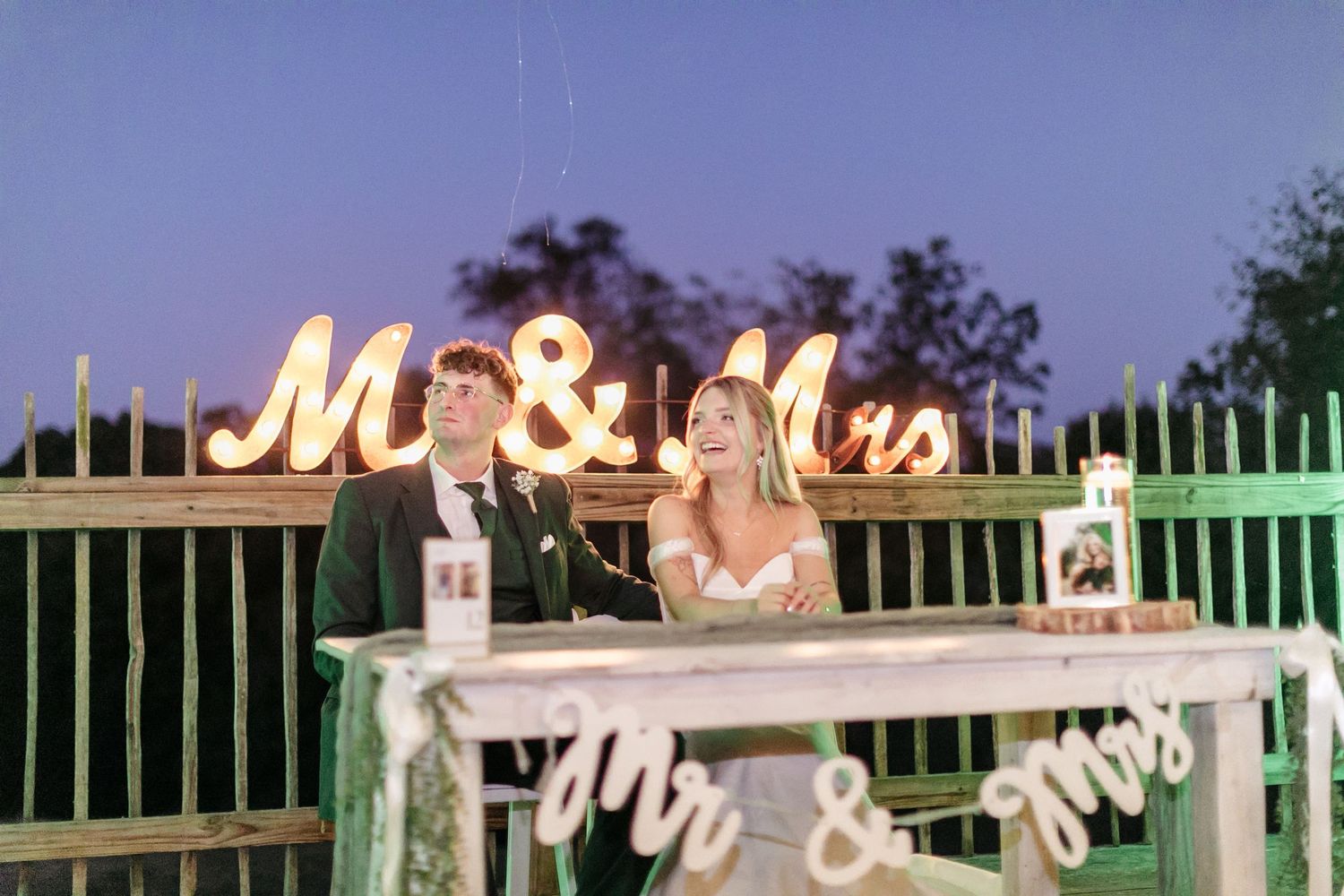 An outdoor wedding reception at dusk with illuminated 'Mr & Mrs' signs and string lights.