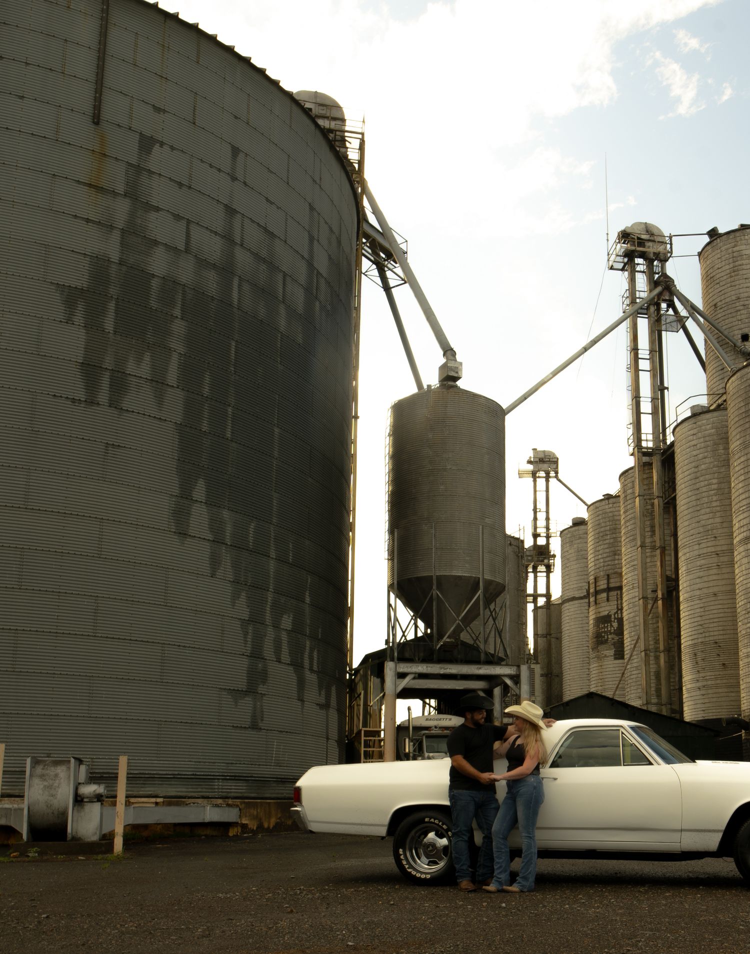Couple standing beside a classic white car near large grain silos at a rural agricultural facility.