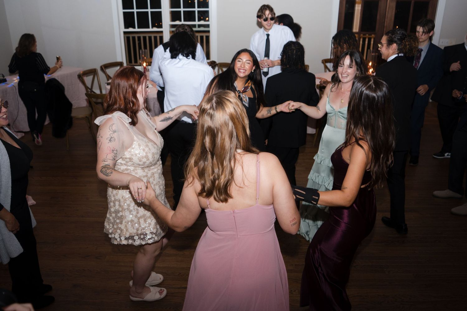 Party guests in formal attire dance together on a wooden floor at an indoor evening celebration.