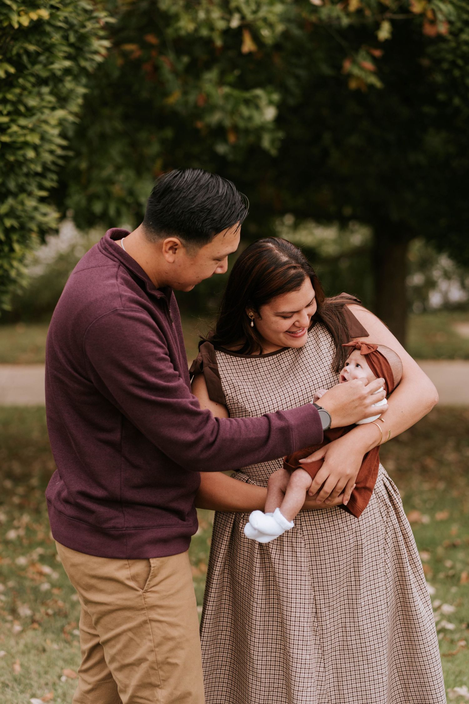 A couple in coordinating burgundy and beige outfits share tender moments with their baby in an outdoor autumn setting.