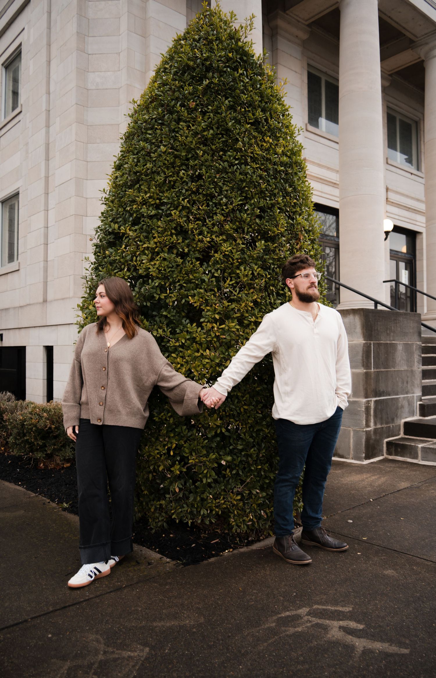 Couple holding hands in front of a tall evergreen shrub outside a stone building.