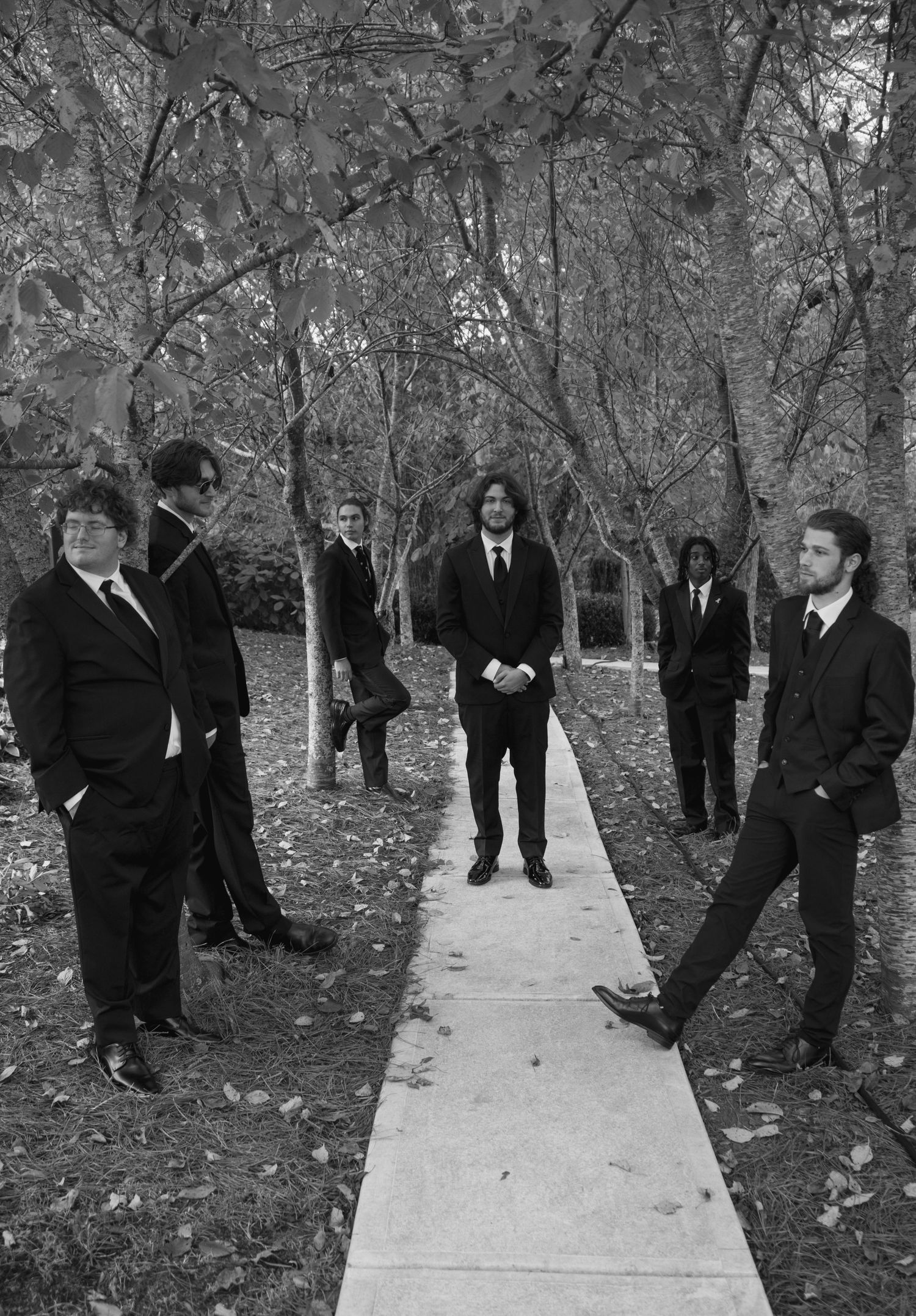 Wedding party members in dark suits playfully pose along a garden path in autumn.