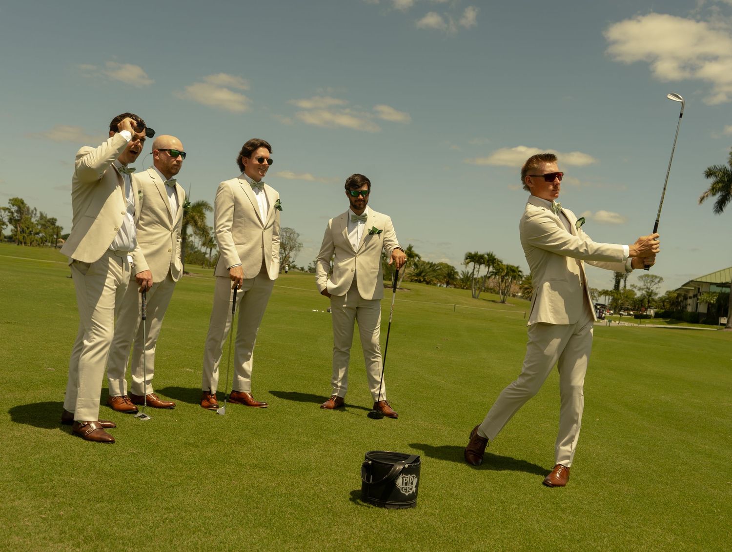 Five groomsmen in white suits playing golf on a sunny green course during a wedding celebration.