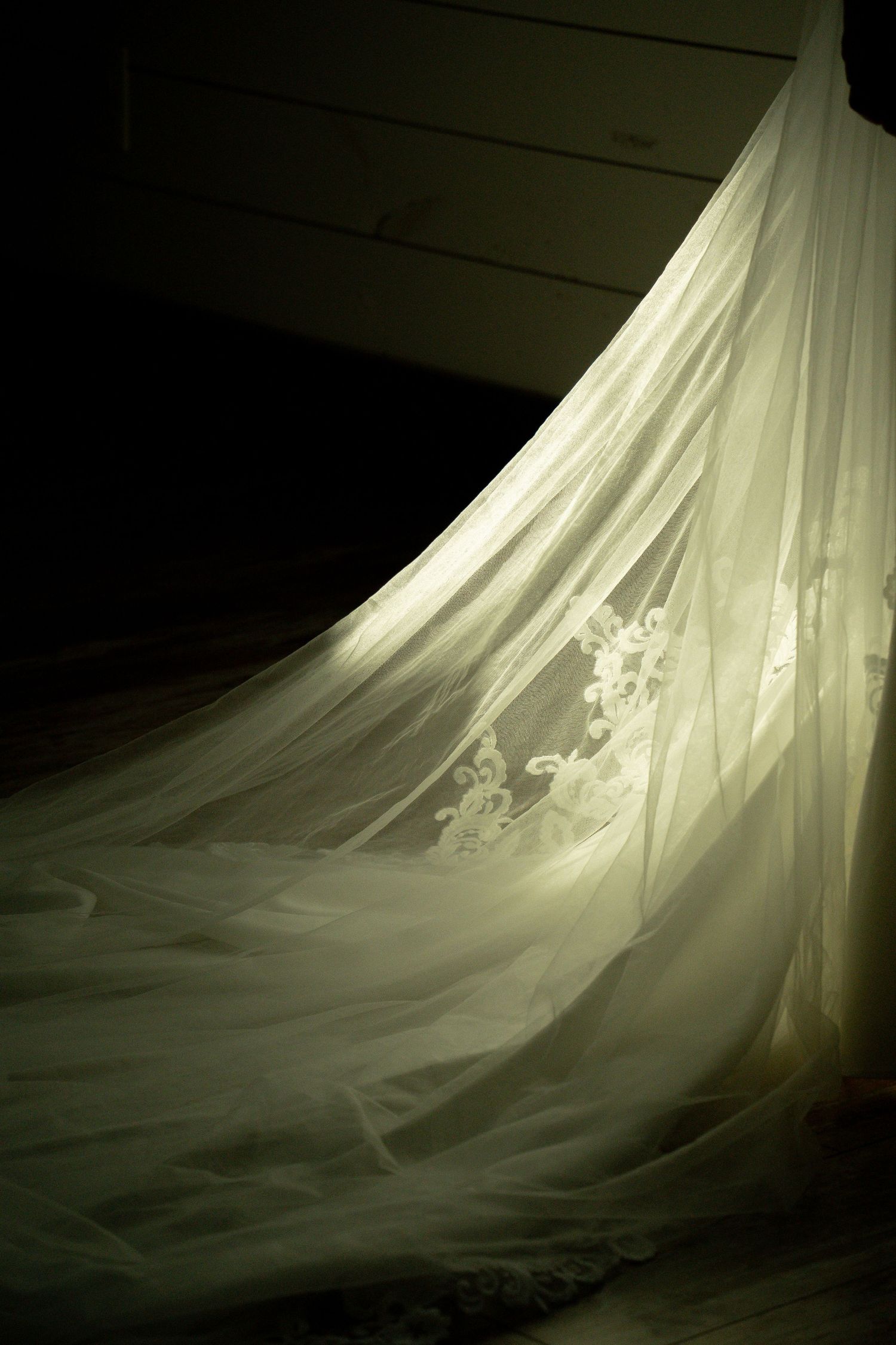 Elegant wedding veil with lace applique detail draped on wooden floor in soft dramatic lighting.