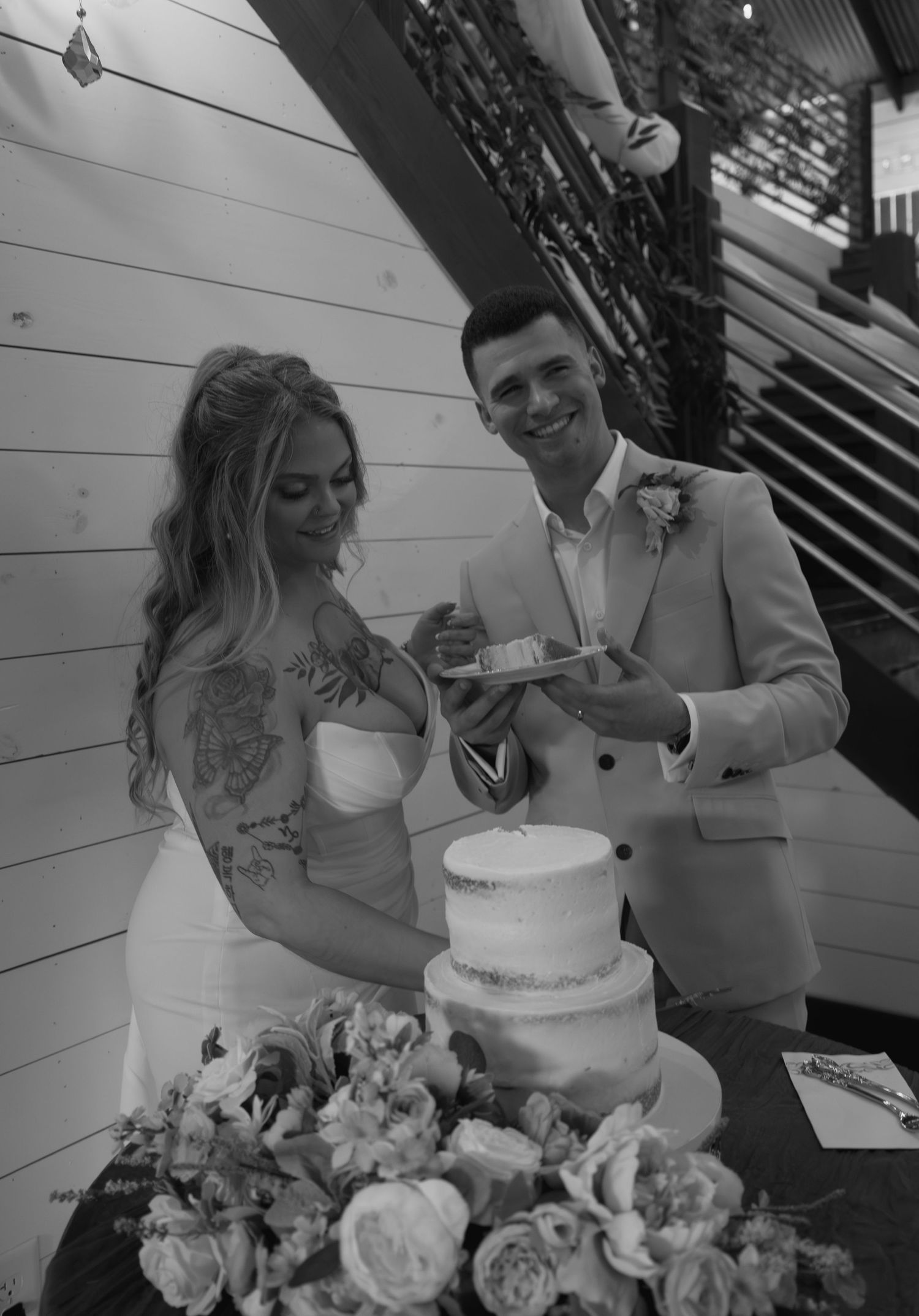 Bride and groom cutting their two-tier wedding cake together, smiling, surrounded by floral arrangements.