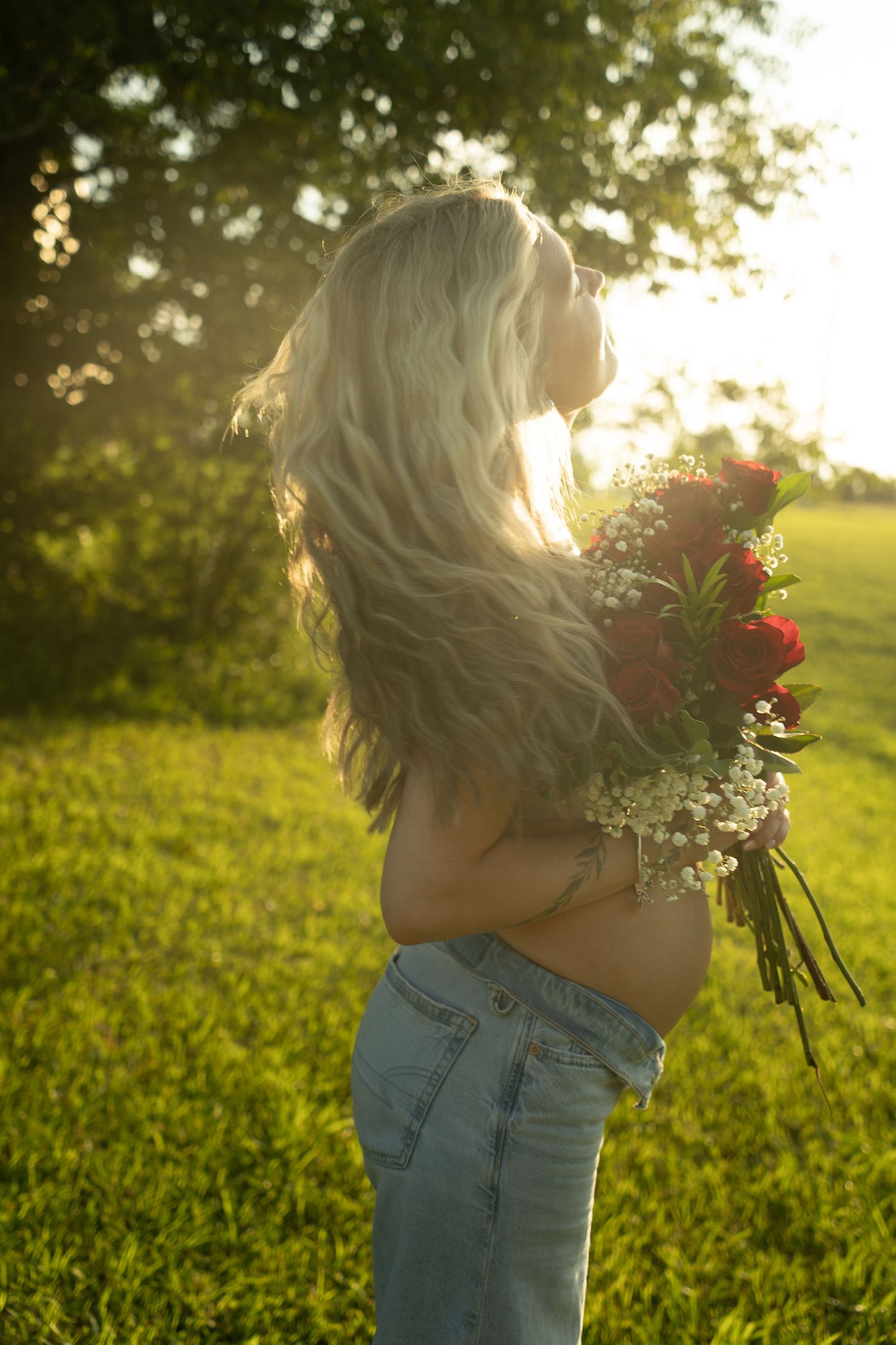 Pregnant woman with long blonde hair holding red roses bouquet in sunny green field wearing jeans.