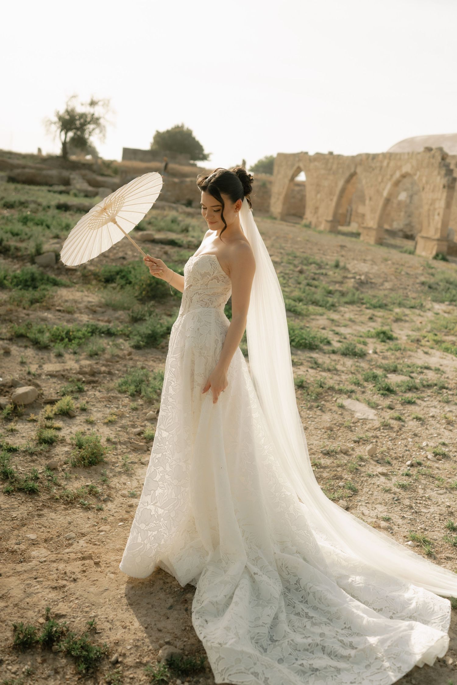 Bride in white lace gown with veil holds parasol umbrella at ancient ruins outdoor wedding photo.