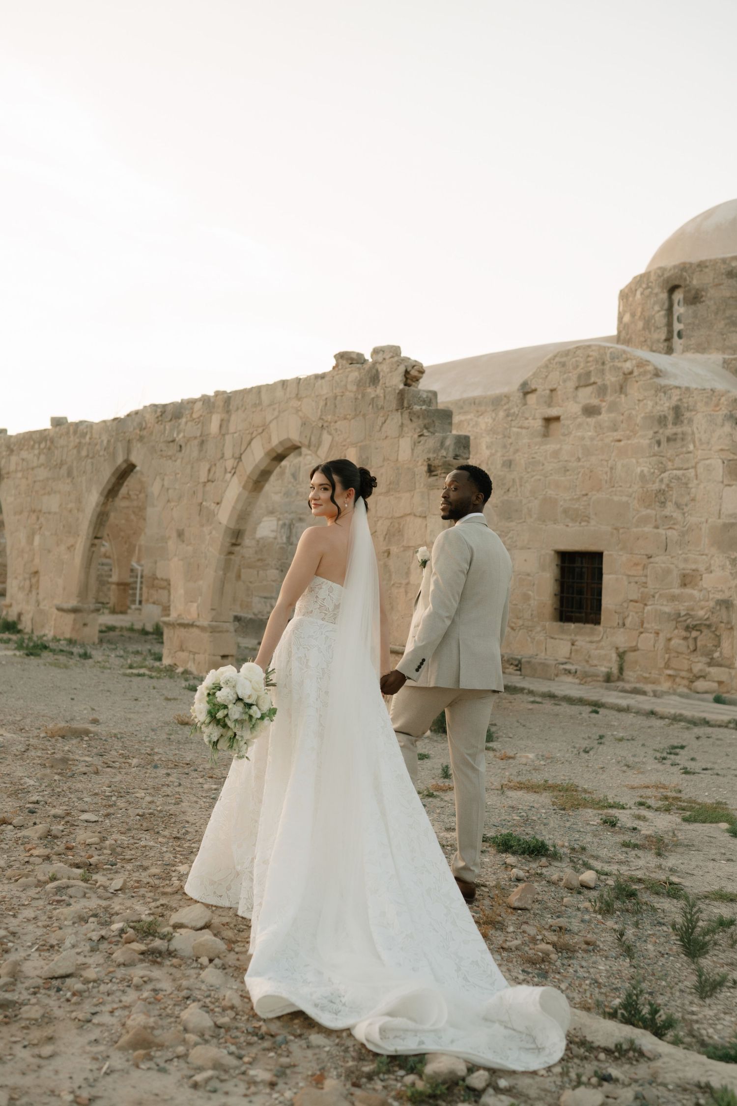 Bride and groom holding hands at ancient stone ruins, bride in elegant white gown with long train holding white bouquet.