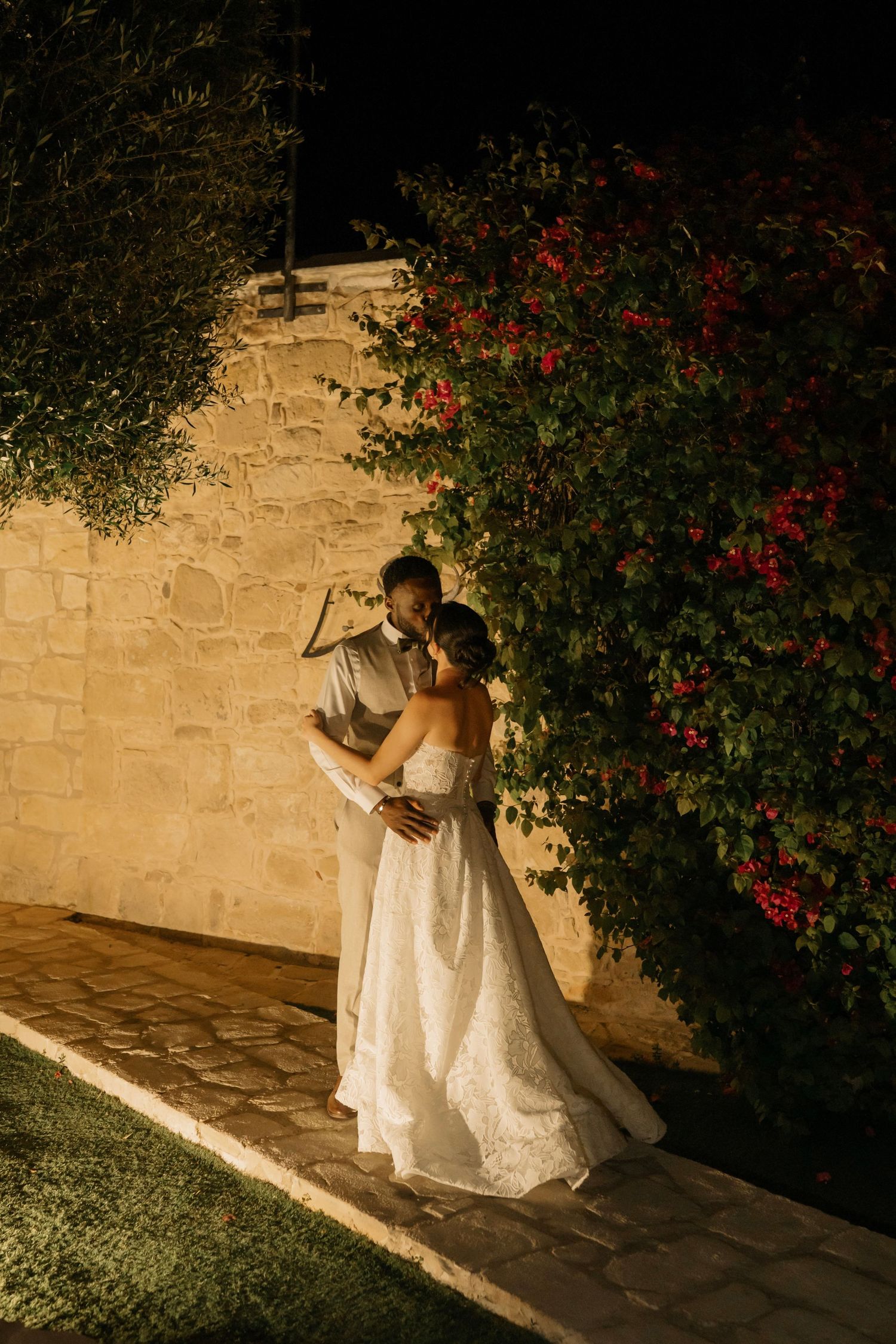 Bride and groom share romantic embrace at night by stone wall with red bougainvillea flowers at outdoor wedding venue.