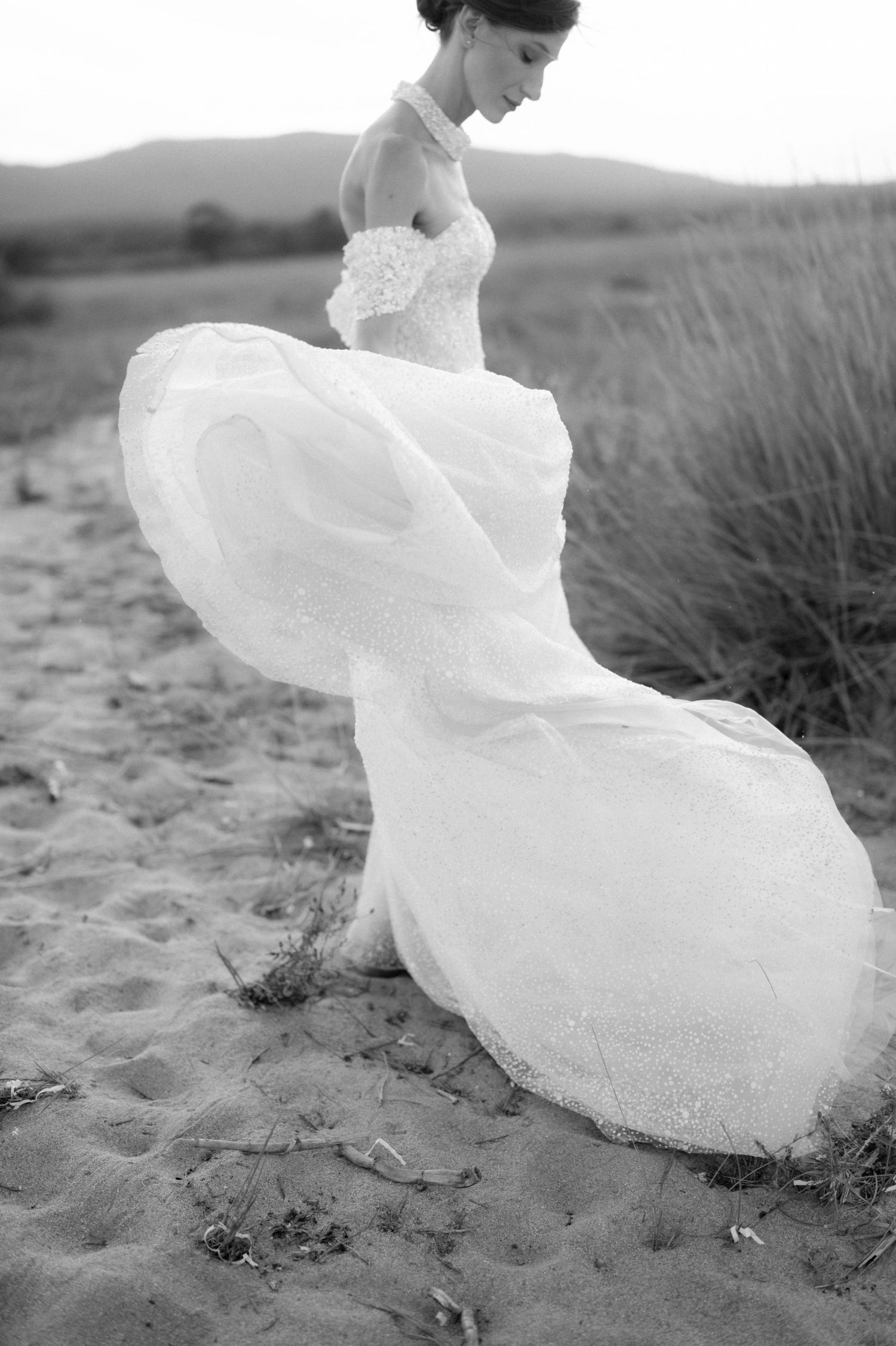 Black and white photo of a bride in a flowing white gown walking on sandy beach, dress billowing in the wind.