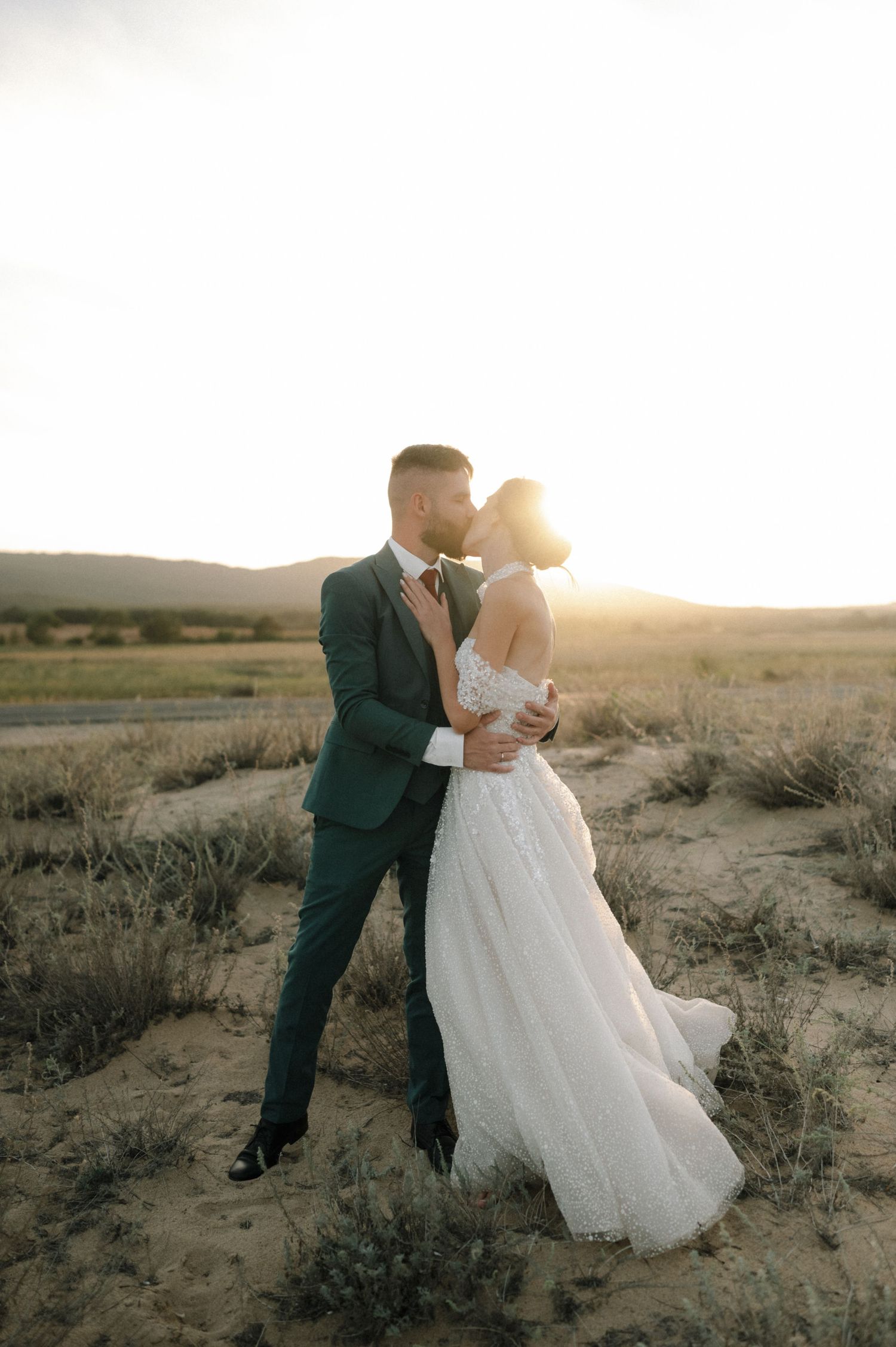 Bride and groom share a sunset kiss in a desert landscape, with golden light glowing behind them.