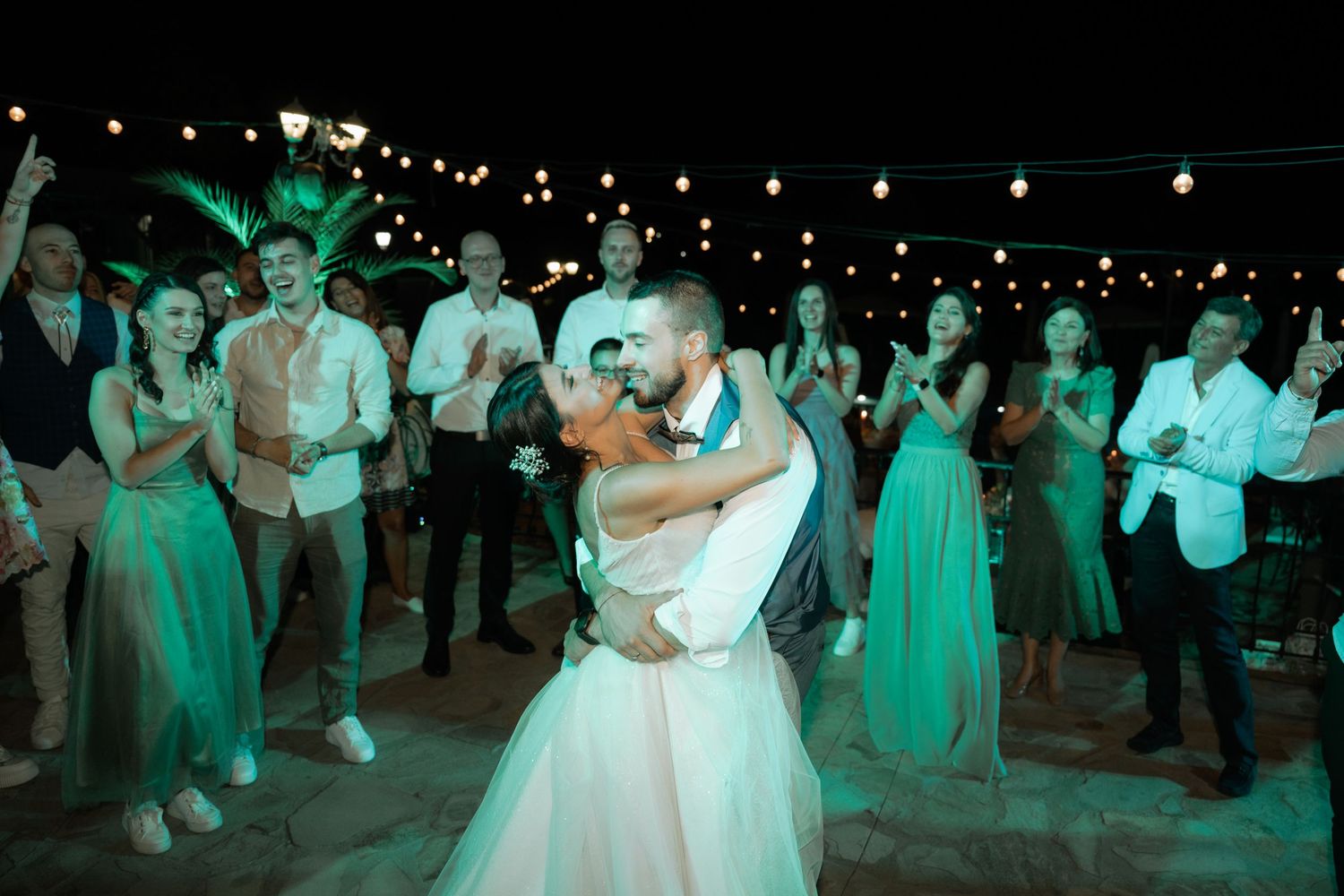 Wedding guests dance together on a dimly lit dance floor illuminated by turquoise lighting at an evening reception.