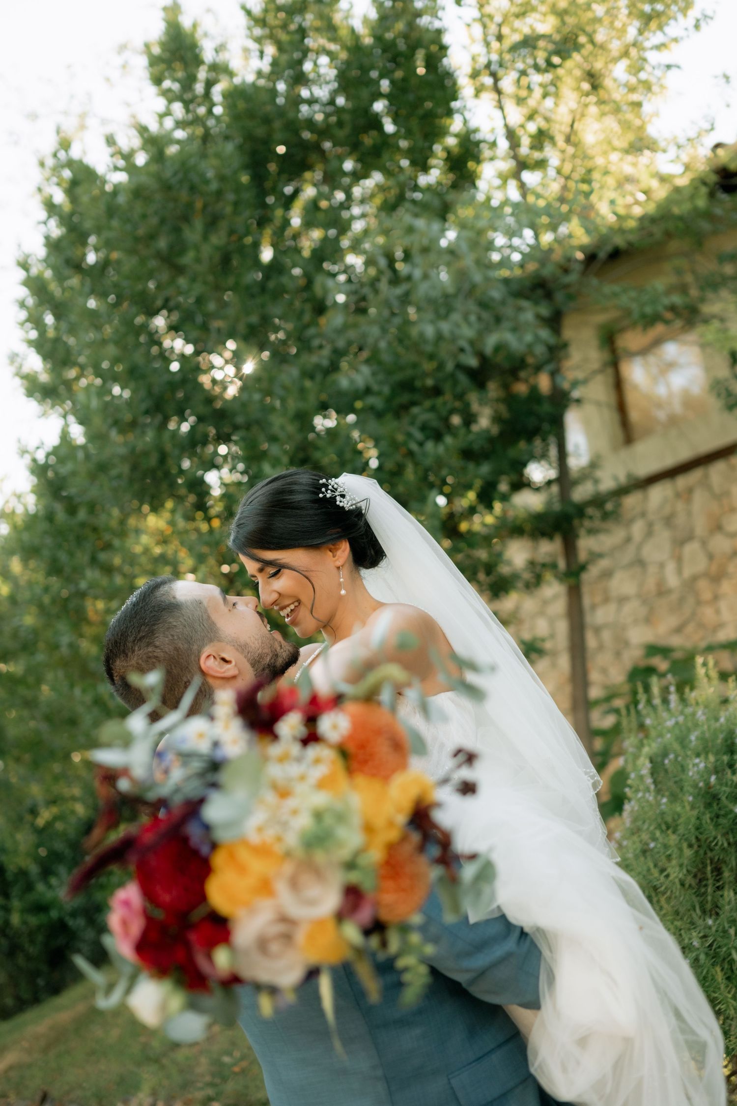 A romantic wedding couple embraces outdoors with vibrant autumn-colored flowers against a backdrop of lush green trees.