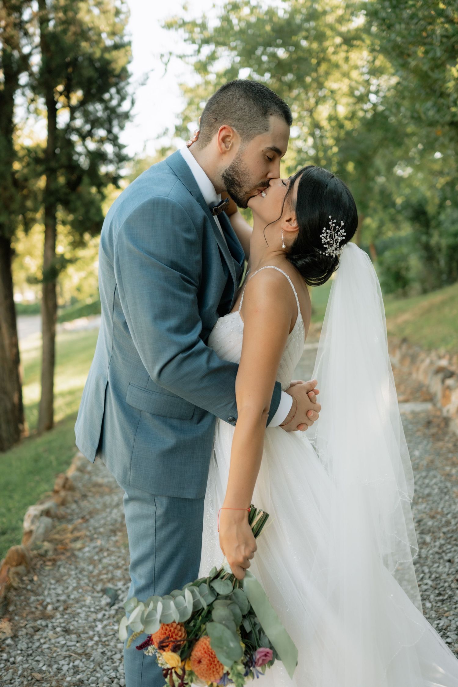 A romantic wedding couple shares intimate kisses on a gravel path surrounded by trees in golden sunlight.