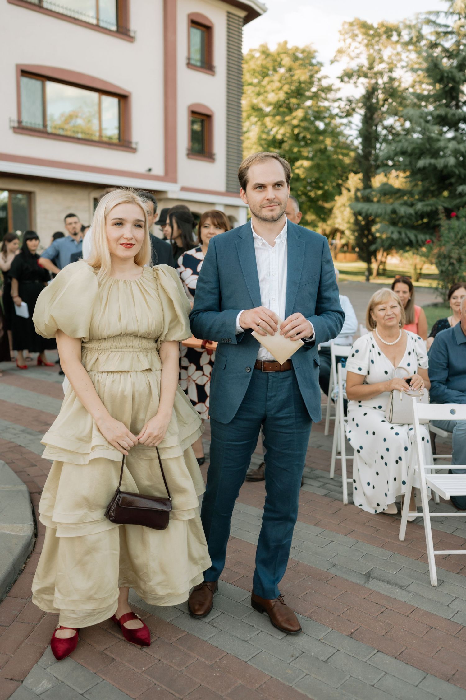 Guests at an outdoor event stand on a brick patio, with a woman in a beige tiered dress and man in a navy suit.