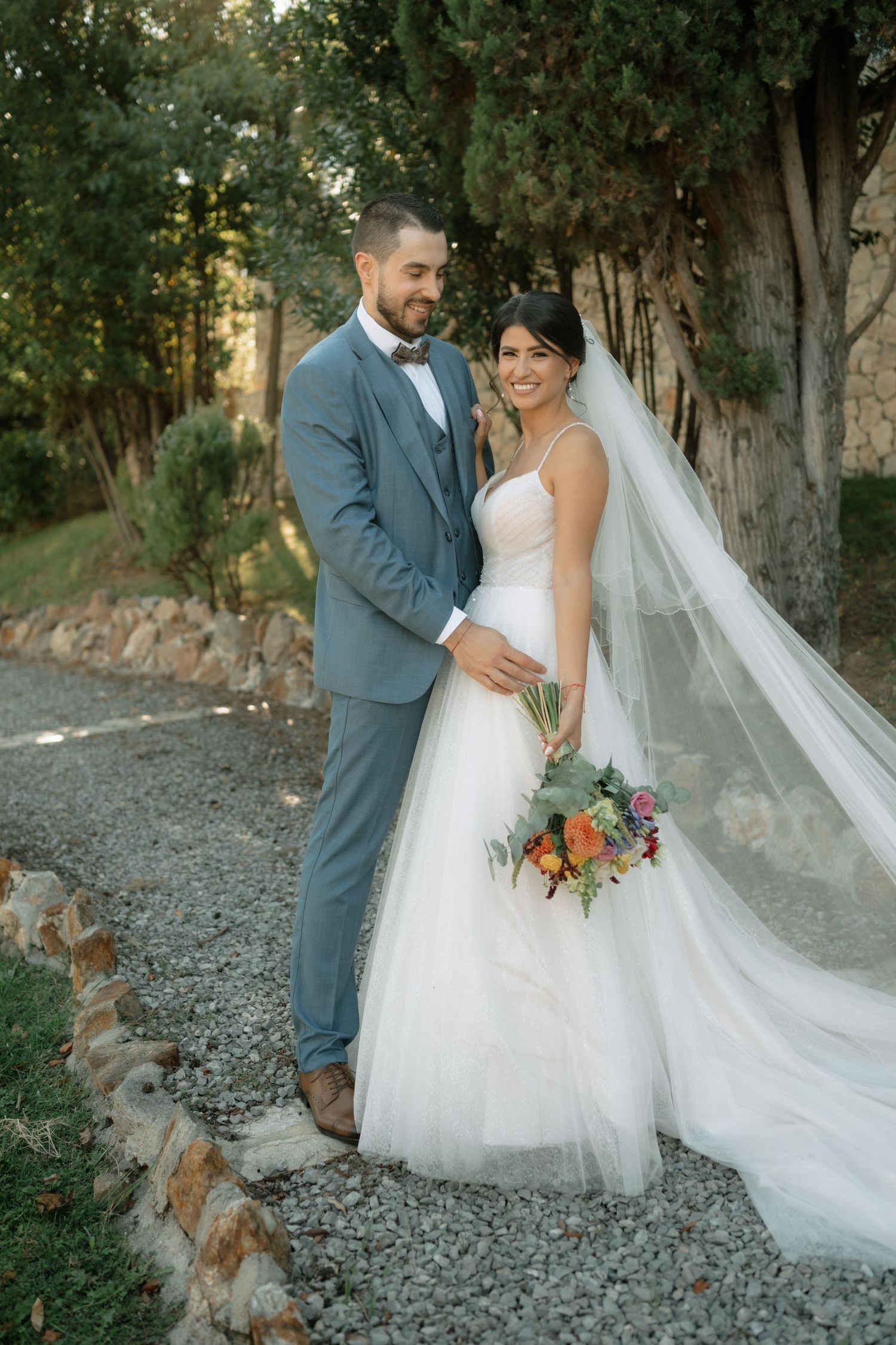 A couple poses together on a gravel path surrounded by trees, with the bride wearing a flowing white gown and veil.