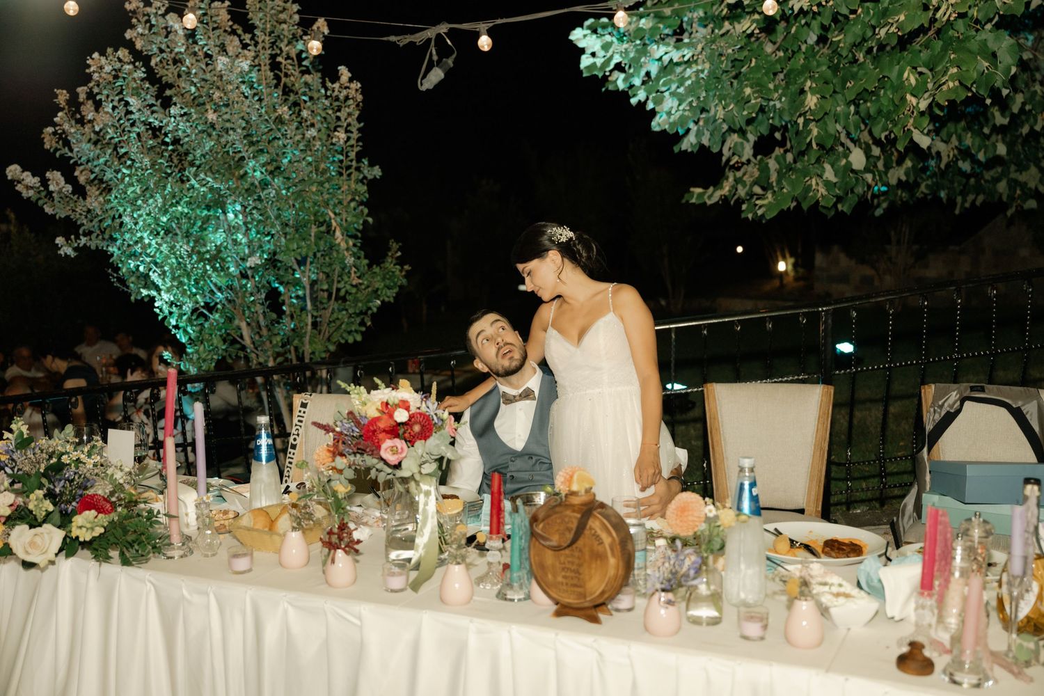 An outdoor evening wedding reception with an elegantly decorated dining table featuring floral arrangements and lanterns.