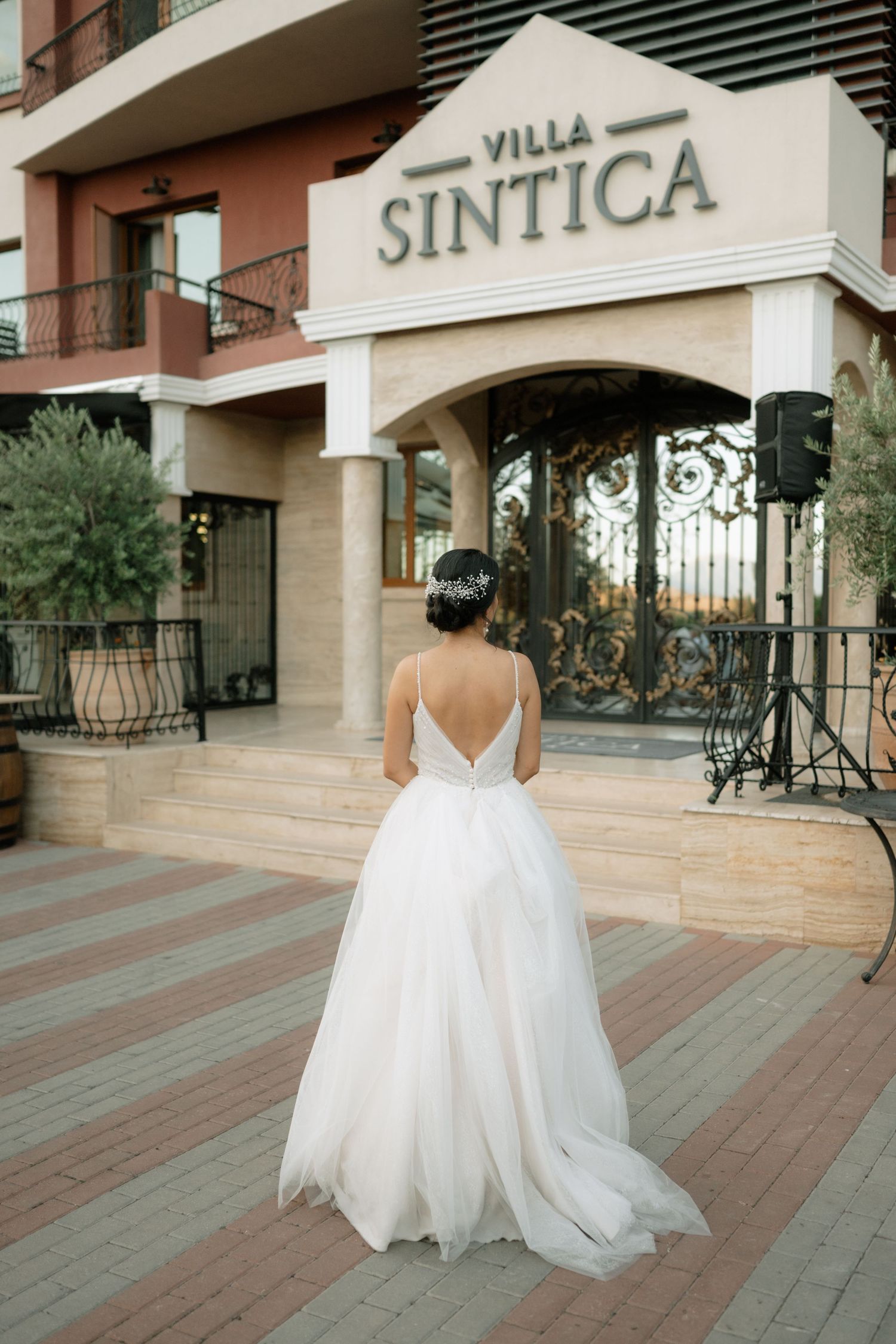 A bride in a flowing white backless gown stands in front of Villa Sintica's elegant entrance.