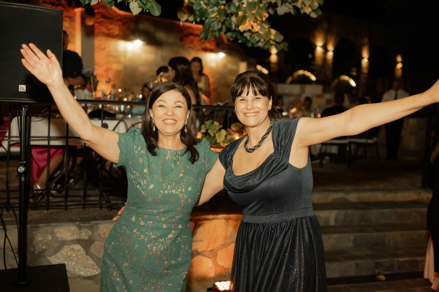 Two people in evening dresses pose with outstretched arms at a nighttime outdoor gathering.