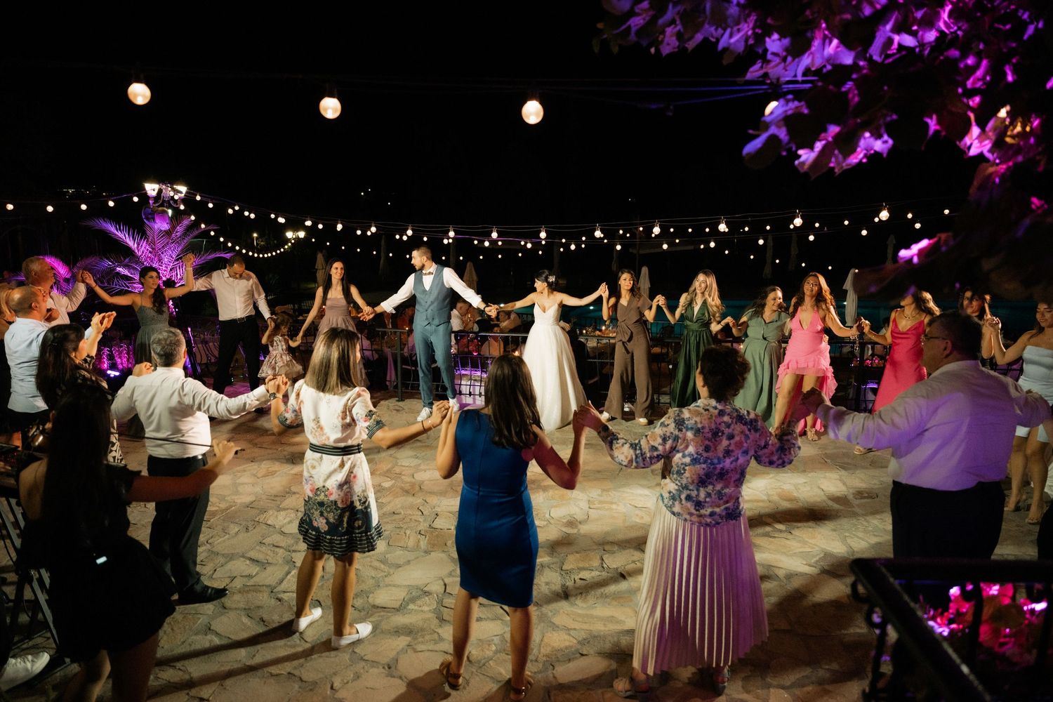 Wedding guests dance under twinkling string lights at an outdoor evening reception with purple decor accents.