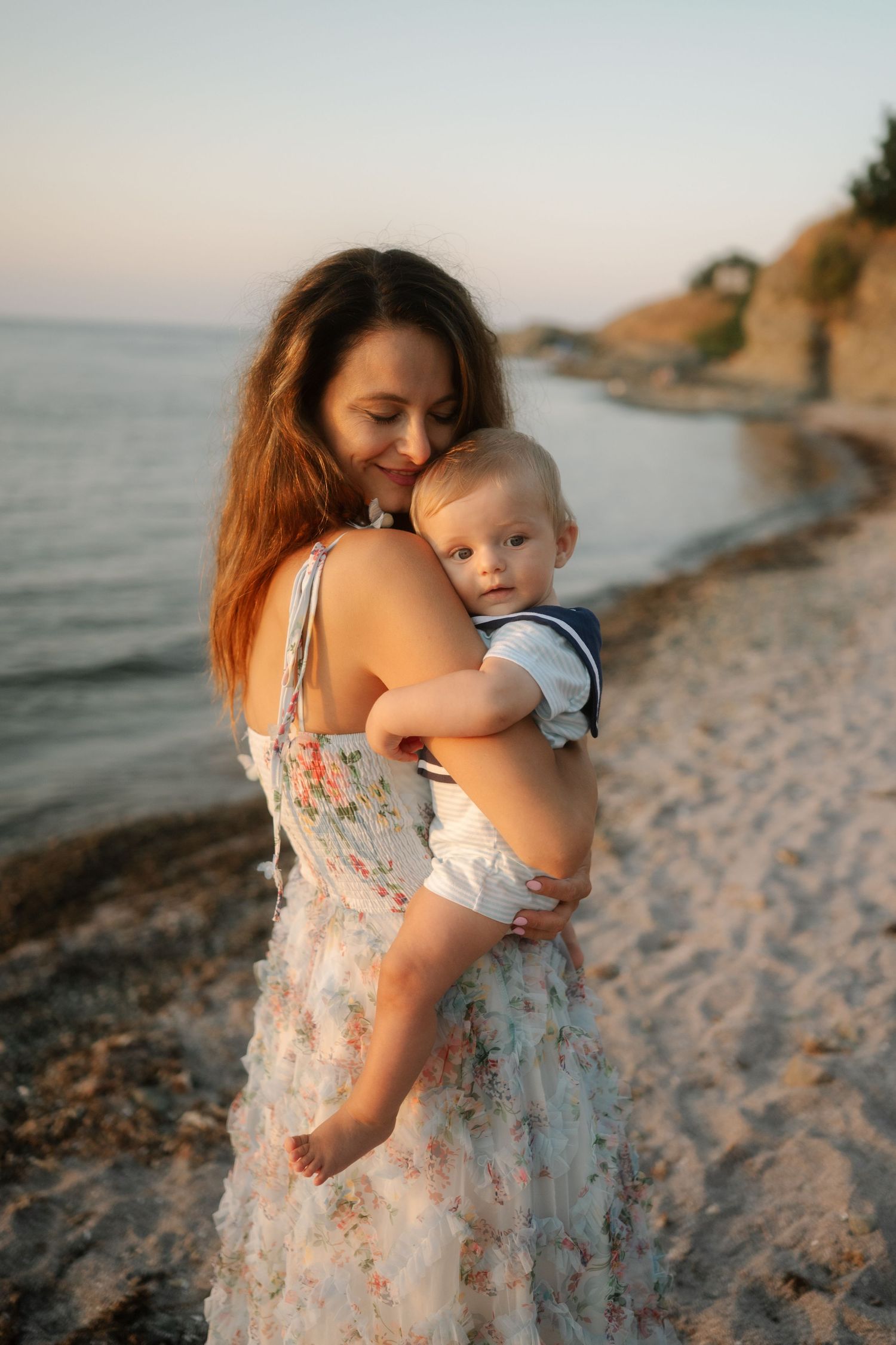 A parent and baby share tender moments on a sandy beach at sunset while wearing floral summer clothing.