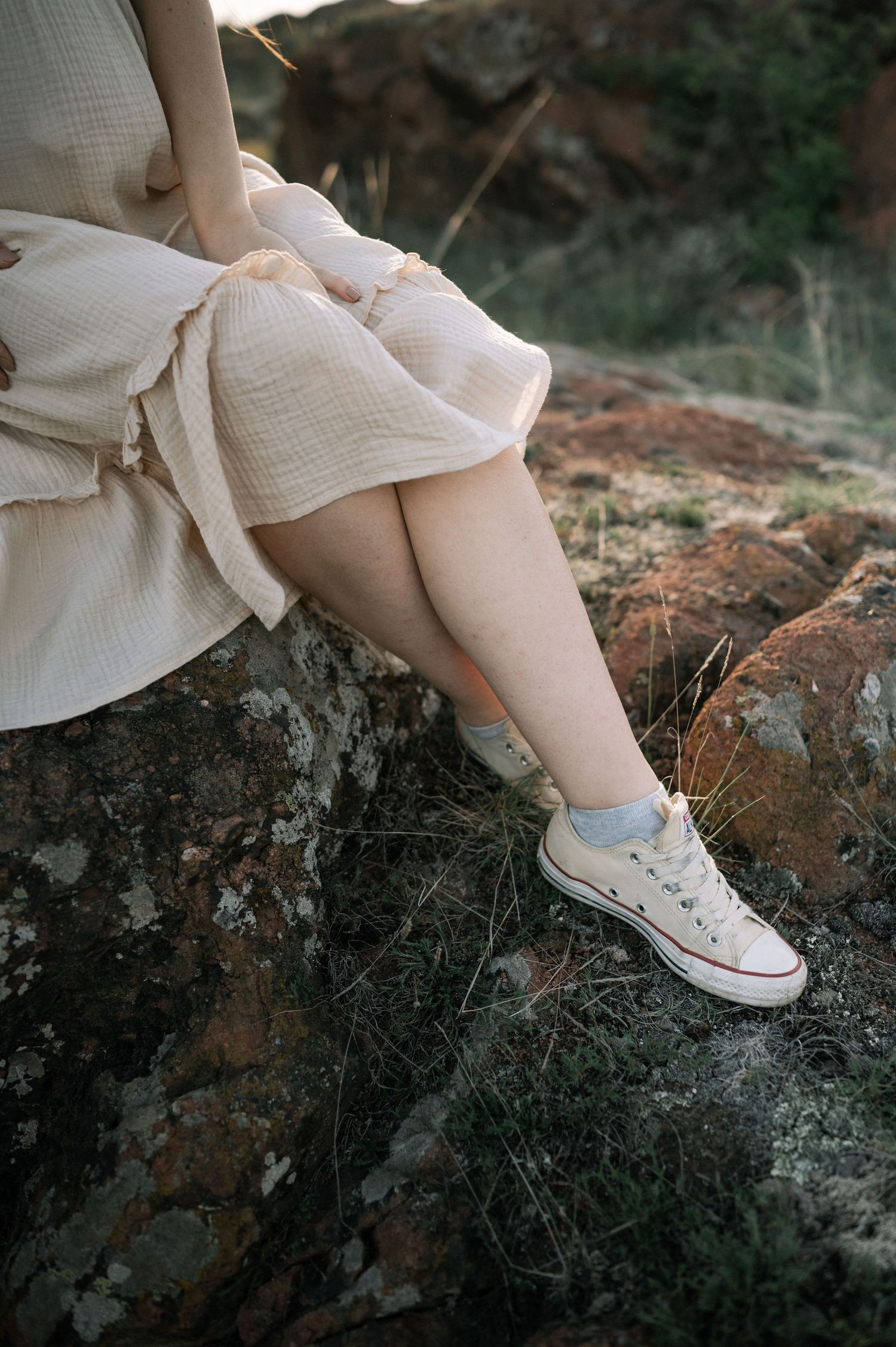 Close-up view of white canvas sneakers and dress hem resting on dark rocks in a moody outdoor setting.