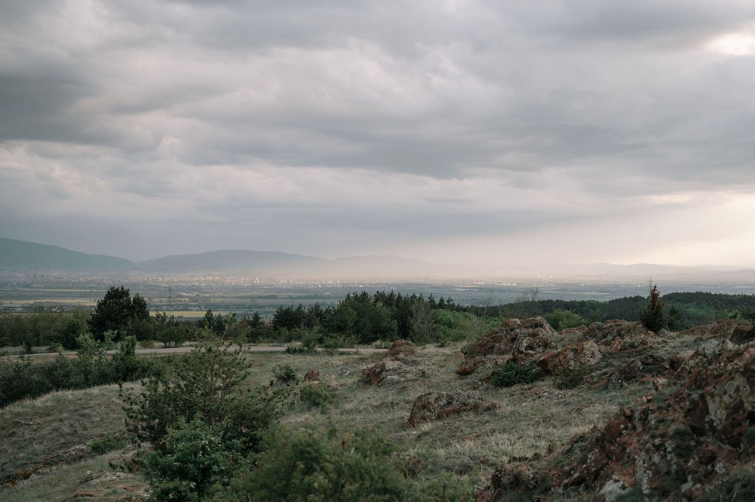 Panoramic view of a rugged mountain landscape with dramatic cloudy skies and scattered vegetation in the distance.