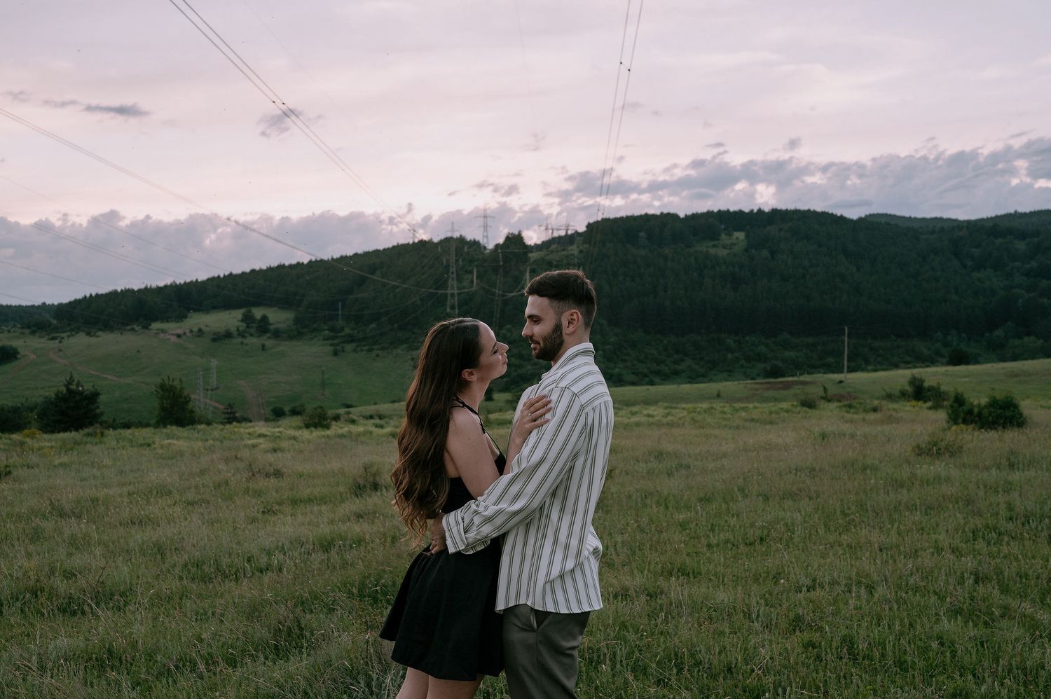 Two people share an intimate moment while standing in a vast meadow with mountains visible in the distance.